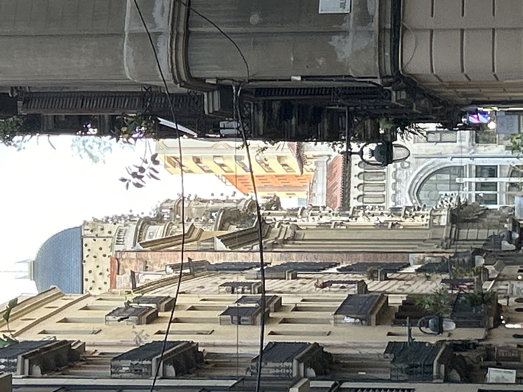 This image captures an urban street scene from an elevated perspective. On the left, a substantial grey architectural structure, possibly an elevated walkway, bridge, or part of a building's underside, extends into the frame. Multiple dark wires and pipes hang down from this structure, and some green foliage emerges from its edges.
To the right and receding into the distance, a cluster of multi-story buildings forms the core of the scene. These buildings exhibit varied architectural styles, featuring numerous windows, balconies, and detailed facades in shades of ochre, beige, grey, and brown. Some rooftops are visible, adorned with various vents and small structures, and a few also host potted plants or small garden areas. A distinctive blue dome caps a building towards the lower right portion of the image. An arched window can be seen on another building in the mid-ground.
The background is dominated by a bright, white sky, indicating it is daytime, though the lighting suggests it might be overcast or hazy. No people or specific activities are visible, presenting a static urban snapshot. Several thin black wires crisscross the entire view, likely utility lines. Faint, unreadable text or labels are present on some elements of the elevated grey structure and other buildings. Given the context, the architecture is consistent with a city like Barcelona, Spain.