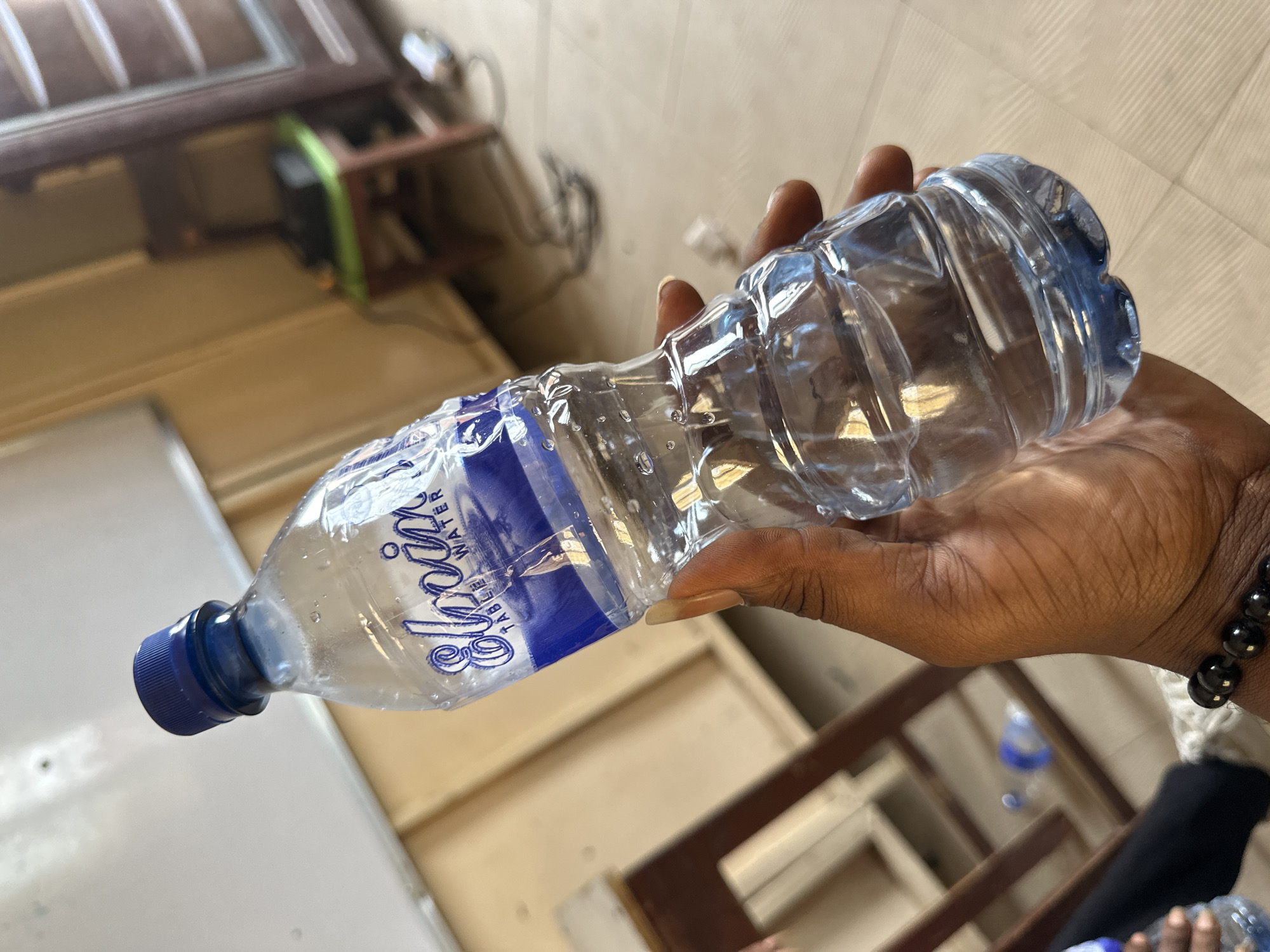 A close-up, slightly tilted photograph captures a person's dark-skinned hand holding a partially full clear plastic water bottle. The hand, positioned in the center-right of the frame, has a prominent light-colored fingernail on one finger and a black beaded bracelet on the wrist. The water bottle features a blue screw cap and a blue and white label that clearly displays the brand name "Sipora" in a stylized script, with "TABLE WATER" written beneath it. Several small water droplets or condensation are visible on the exterior of the bottle, suggesting it might be cold or recently handled.
The background is intentionally blurred, indicating an indoor setting. Faint details suggest light-colored walls or surfaces and some brown wooden structures, possibly furniture or a door frame. A small, blurry green object is also discernible in the background. The bright lighting in the image suggests either natural daylight entering indoors or ample artificial lighting. The overall scene shows a simple, everyday act of holding a water bottle. The context provided indicates this scene is set in Lagos, Nigeria.
