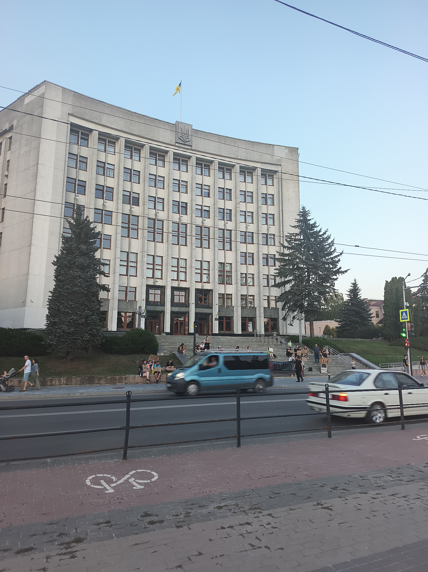 The image captures a wide shot of the Ternopil City Council building in Ternopil, Ukraine. The building, a large, imposing structure with multiple windows and a prominent coat of arms at the top, dominates the background. In front of the building, a set of wide stairs leads up to the entrance. On the stairs and the surrounding grassy area, a group of people are gathered. Some are holding signs, suggesting a demonstration or protest.
In the foreground, a street runs parallel to the building, with a light blue van and a white car in motion, indicated by motion blur. A bicycle lane is marked on the pedestrian area to the right of the street. The overall environment suggests a public space in a city, likely during daylight hours given the clear sky. Electrical wires crisscross the sky above the building and the street, common in urban settings.