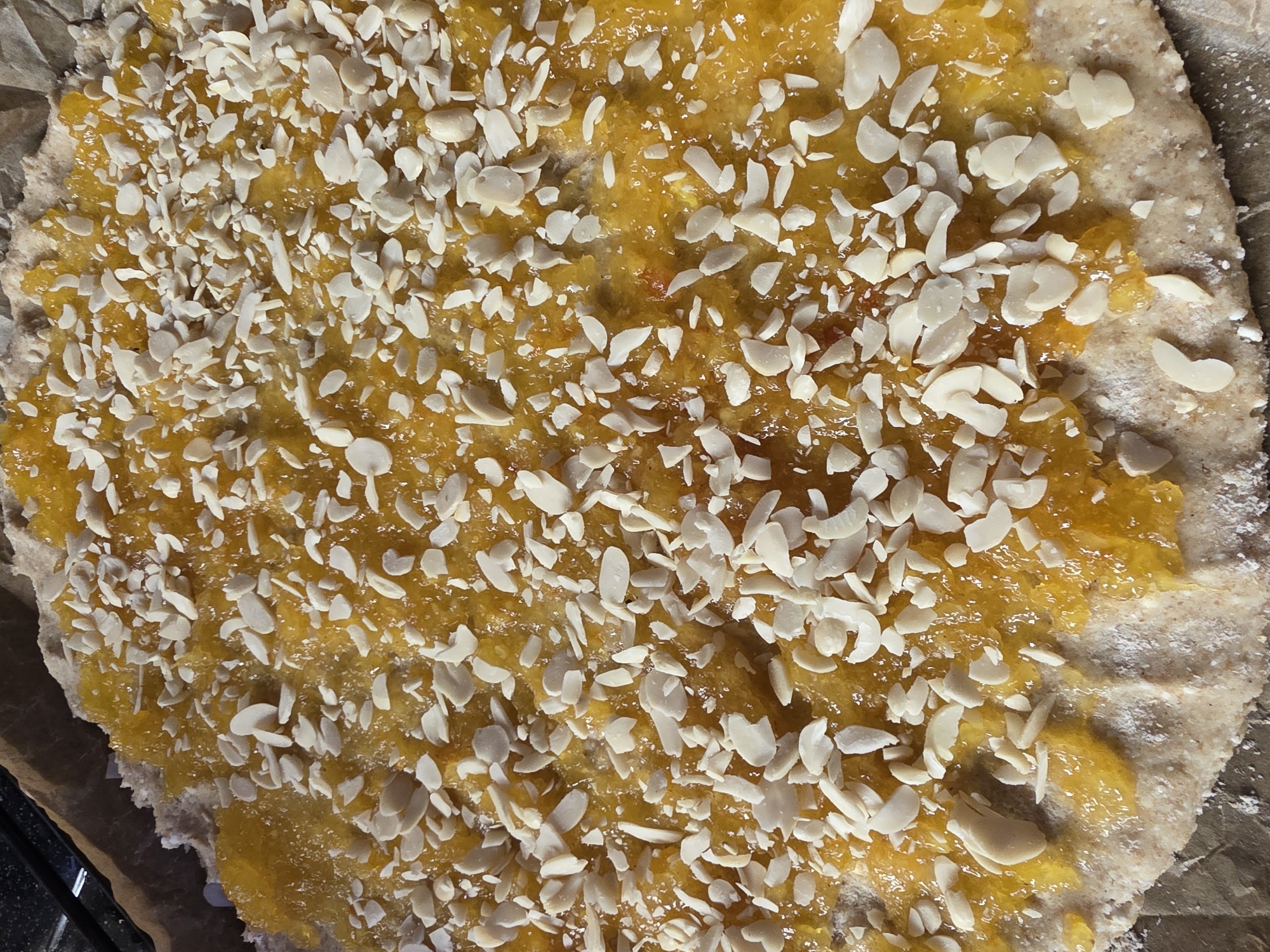 The image shows a close-up, top-down view of a raw, circular pastry topped with a vibrant orange filling and sprinkled with sliced almonds. The pastry base appears to be made of whole wheat flour, with a slightly uneven, rustic texture. The orange filling is chunky and glistening, suggesting it could be fruit jam or preserve. The sliced almonds are scattered unevenly across the filling, with some pieces overlapping. The pastry is resting on a sheet of brown parchment paper, which is placed on a dark, possibly a baking sheet. The lighting is bright, casting some subtle shadows and highlighting the textures of the ingredients. There is no indication of the city of Saint Petersburg, Russia in the image.