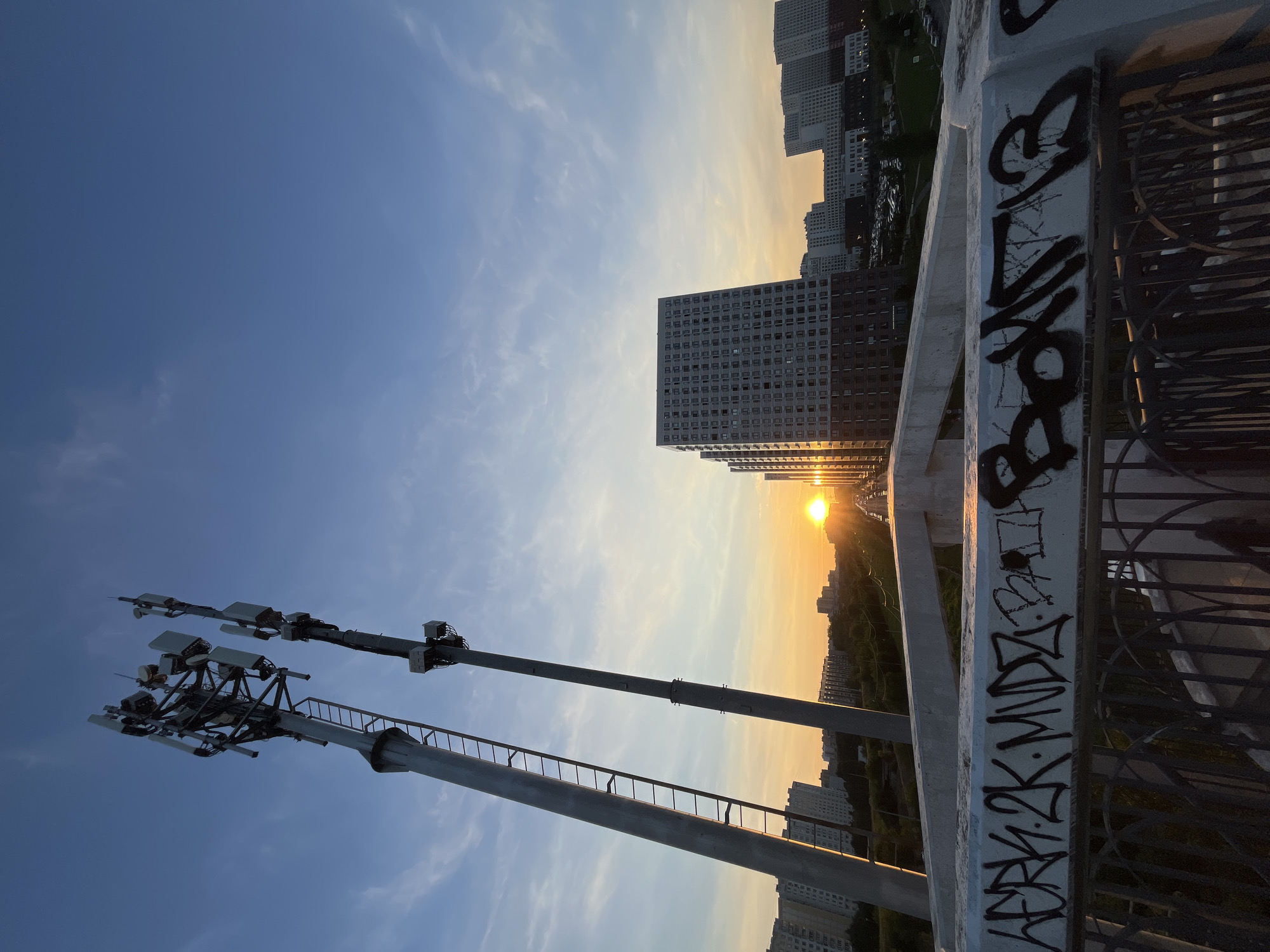 The image depicts a wide-angle view of an urban landscape at sunset. Two tall telecommunication towers dominate the left side of the frame, silhouetted against a clear blue sky with wispy clouds. The sun, a bright orb, is setting on the horizon, casting a warm orange glow across the sky and illuminating several high-rise apartment buildings in the background. The foreground features a concrete structure, possibly a bridge or overpass, with visible graffiti. The graffiti on the concrete barrier is in black spray paint and appears to be stylized lettering. Below this barrier, a dark metal railing is visible. The overall impression is of a modern city at dusk, with technology, residential structures, and natural light creating a striking contrast.