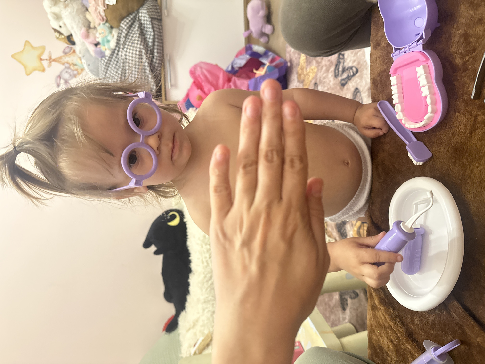A child with light brown hair styled in a small ponytail wears purple-rimmed glasses. The child is partially undressed, exposing the upper torso, and appears to be interacting with a dental kit toy. A partially visible hand is raised in the foreground, partially obstructing the view of the child.
The dental kit includes a purple toothbrush, a set of artificial teeth in a purple container, and other dental tools on a white plate. These items are arranged on a brown, textured surface. In the background, there's a wall painted in a light color, adorned with a star-shaped decoration and a dark, plush toy resembling a creature. Other toys and household items are visible in the cluttered background.
The image provides no information about Kharnavy, Russia.
