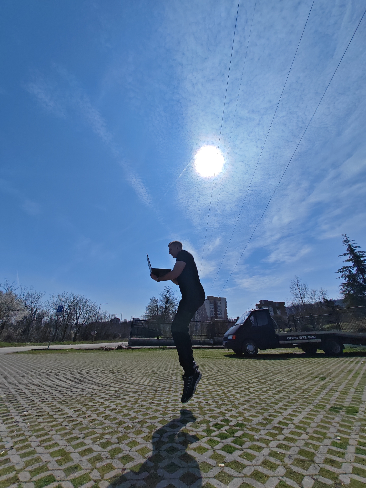 The scene depicts an outdoor environment in Shumen, Bulgaria, under a clear blue sky with scattered wispy clouds. A single adult male, dressed in a dark short-sleeved shirt, dark trousers, and dark athletic shoes, is captured in mid-air with both feet elevated above the ground. He holds an open, dark-colored laptop computer in both hands, positioned in front of him with the screen facing away from the camera. The sun, a brilliant white disc, is prominently visible in the upper-left sky, casting a distinct shadow of the man onto the ground directly below him. The ground surface consists of a grid-patterned concrete paver system with grass growing within its interstices. In the mid-background, a dark flatbed truck is parked, displaying the white text "0896 972 982" on its side. Further in the background, to the left and behind the truck, multi-story residential buildings are visible. Bare deciduous trees, some with white blossoms on the far left, and a green conifer on the far right, line the background. A blue parking sign (P) is visible on a pole in the left-mid background. Multiple parallel overhead power lines stretch across the upper portion of the sky.