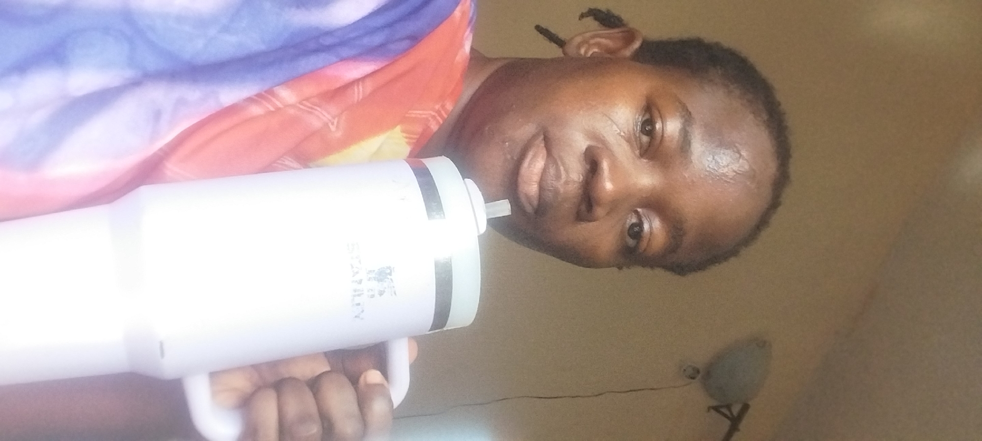 A close-up, selfie-style photo captures a dark-skinned person in Onitsha, Nigeria, holding a large, white insulated tumbler with a handle and straw. The person is wearing a vibrantly colored garment, possibly a wrapper or scarf, with shades of pink, orange, yellow, and blue. Their hair is styled in braids, with one braid visible on the left side of their head. The background is a plain, light-colored wall, and a light fixture is visible in the upper right corner of the image. The tumbler has the "STANLEY" logo visible on its side, along with some other text that is partially obscured. The person's left hand is holding the handle of the tumbler, and their right hand is out of frame. The lighting is somewhat diffused, suggesting indoor conditions.