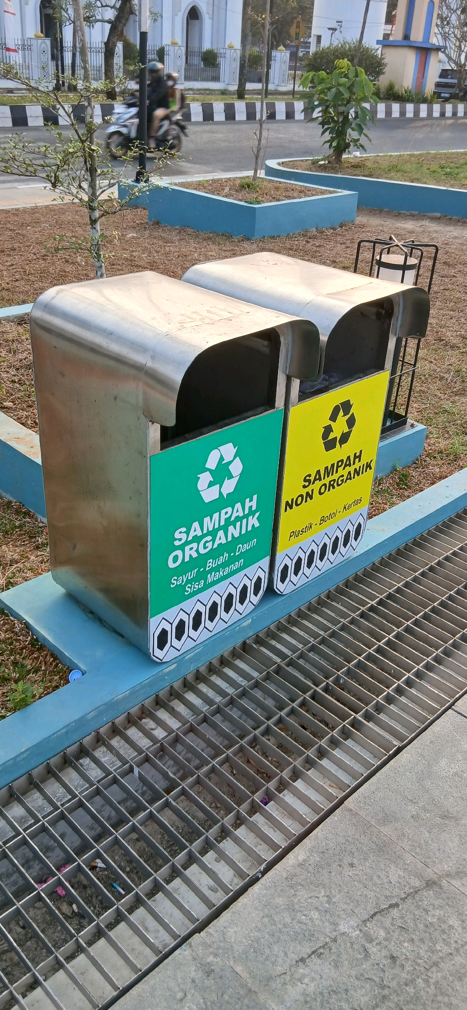 This is a medium shot, eye-level, outdoor photograph of two stainless steel trash receptacles situated in a park setting in Samarinda, Indonesia. The receptacles are side-by-side, with the left one displaying a green label and the right one a yellow label. Both labels feature a recycling symbol. The green label reads "SAMPAH ORGANIK" with smaller text below indicating "Sayur - Buah - Daun Sisa Makanan". The yellow label reads "SAMPAH NON ORGANIK" with smaller text below indicating "Plastik - Botol - Kertas". The trash cans are elevated on a blue concrete structure. In front of the trash cans, a metal grate covers a drainage ditch. In the background, a paved road is visible with a black and white striped curb. A motorcycle with two riders is blurred in motion on the road. Beyond the road, there are trees, some with sparse foliage, and a building with white walls and arched windows. The ground around the trash receptacles is covered with dry grass and mulch. The overall lighting suggests it is daytime, possibly late afternoon.