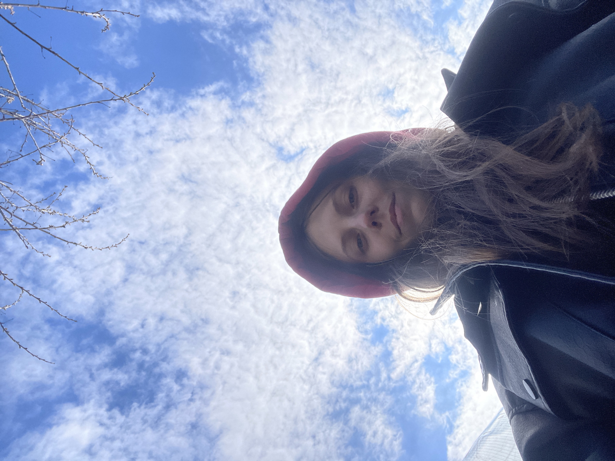 A low-angle photograph of a person wearing a red hooded sweatshirt and a dark jacket. The person's face is visible, and they are looking down at the camera. The background consists of a blue sky with scattered white clouds and bare tree branches in the upper left corner. The location is likely Oktyabrsky, Russia, though this cannot be definitively confirmed from the image alone.