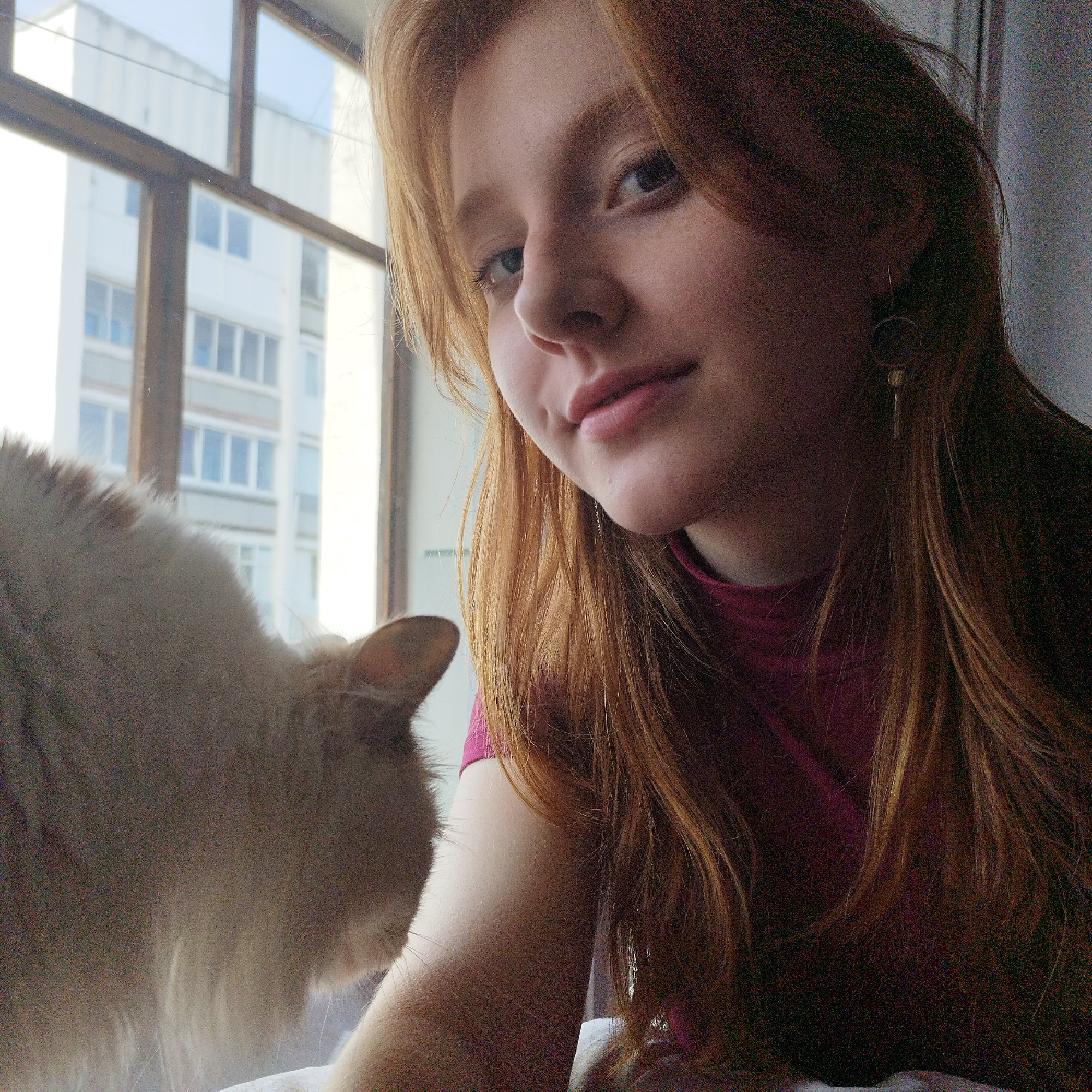A selfie depicts a young woman with long, reddish-blonde hair, wearing a pink top, positioned indoors near a window. Her face is slightly angled towards the camera with a subtle smile. She has fair skin and light-colored eyes. A cat, predominantly white, is partially visible on the left side of the frame, near the window. Outside the window, a tall, multi-story building is seen, suggesting an urban setting. The scene appears to be taken during daylight hours due to the natural light filtering through the window. The location may be in Yekaterinburg, Russia, based on contextual information, although this is not explicitly confirmed by the image alone. The indoor setting suggests a domestic environment. There are window frames visible and a bright building viewable.