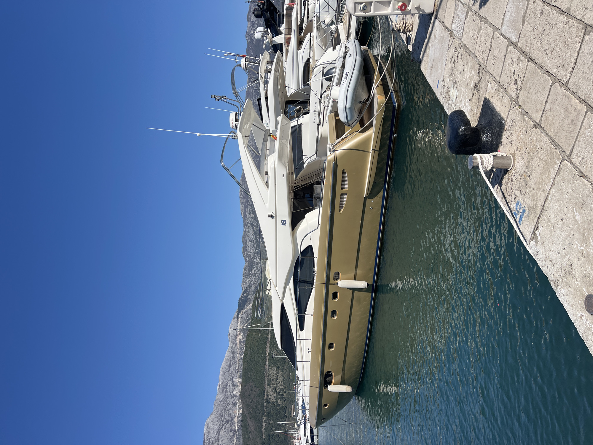 The image shows a docked yacht in Budva, Montenegro, with a clear blue sky above and mountains in the background. The yacht is predominantly white and gold, with multiple decks and a smaller inflatable boat attached. The yacht is moored alongside a stone quay. There is a black mooring bollard and white ropes securing the yacht to the quay. The water surrounding the boat is a greenish-blue color, with slight ripples on the surface. Other boats are visible in the background.