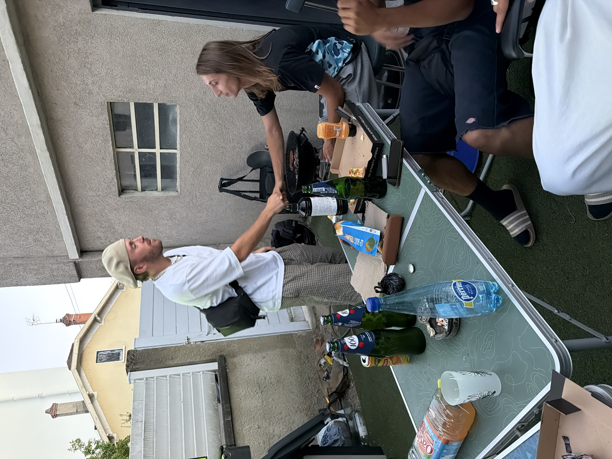 This media file captures a casual outdoor gathering, possibly a backyard barbecue or a relaxed get-together. The scene features at least three people, two of whom are prominently visible. In the foreground, a man wearing a white t-shirt and a tan cap is looking upwards with his eyes closed, seemingly enjoying a moment of leisure. He has a crossbody bag slung over his shoulder and is leaning against a table. To his right, a woman with long brown hair, wearing a black t-shirt with a blue and white camouflage pattern, is leaning over the same table. She appears to be reaching for something or interacting with an object on the table. The table is laden with various items, including several bottles of drinks, a pizza box with visible slices, and other miscellaneous objects. One bottle of "Perrier" water is clearly labeled. The background suggests a residential setting with part of a building visible, including a window with a metal grate. The overall impression is one of a sunny day, given the lighting, and a relaxed, convivial atmosphere. The image is oriented vertically, with the subjects appearing sideways to the viewer. There is also some visible text on the pizza box, reading "LE GOUT ORIGINAL" and "TASTY PIZZA".