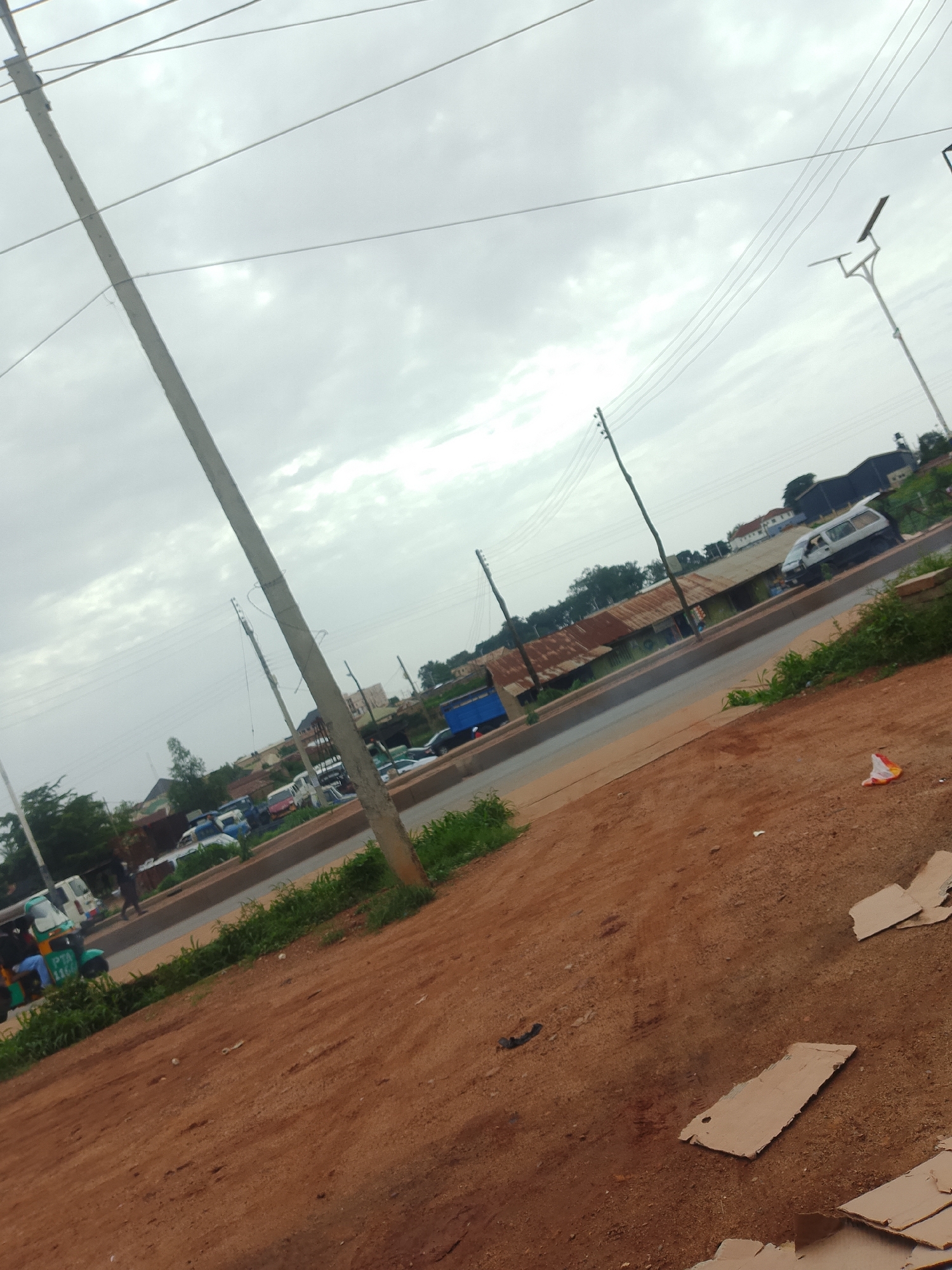 The image shows a street scene in Bukuru, Nigeria. The sky is overcast, suggesting a cloudy or possibly rainy day. A tall utility pole with multiple wires dominates the left side of the frame, leaning slightly. In the background, there is a road with several vehicles, including vans, cars, and a three-wheeled auto-rickshaw. The buildings lining the road appear to be simple structures, some with corrugated metal roofs. There are some green plants and grassy areas along the roadside. The foreground is a patch of reddish-brown dirt with several pieces of cardboard scattered on it.
In the lower left of the image, a three-wheeled auto-rickshaw with passengers is visible. A person is walking on the side of the road in the middle distance. The overall impression is of a busy, yet somewhat undeveloped, urban or semi-urban environment. The time of day is unclear due to the overcast sky, but it appears to be daytime. There is no discernible text in the image. The scene evokes a sense of everyday life and activity.