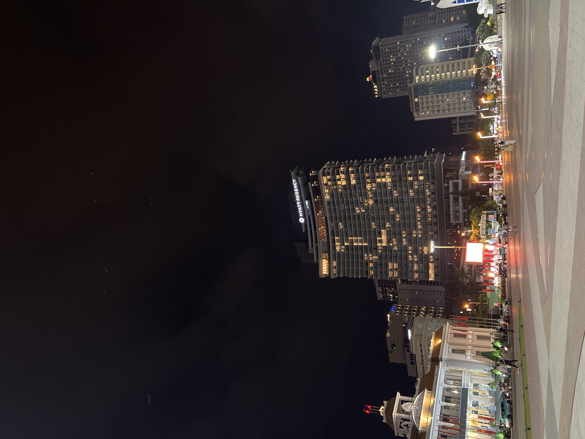 The image shows a nighttime view of Nha Trang Ward, Vietnam. In the foreground are the lighted buildings of the city. The Hyatt Regency is visible in the image. The dark sky fills the background. A light-colored walkway or plaza appears in the foreground right.