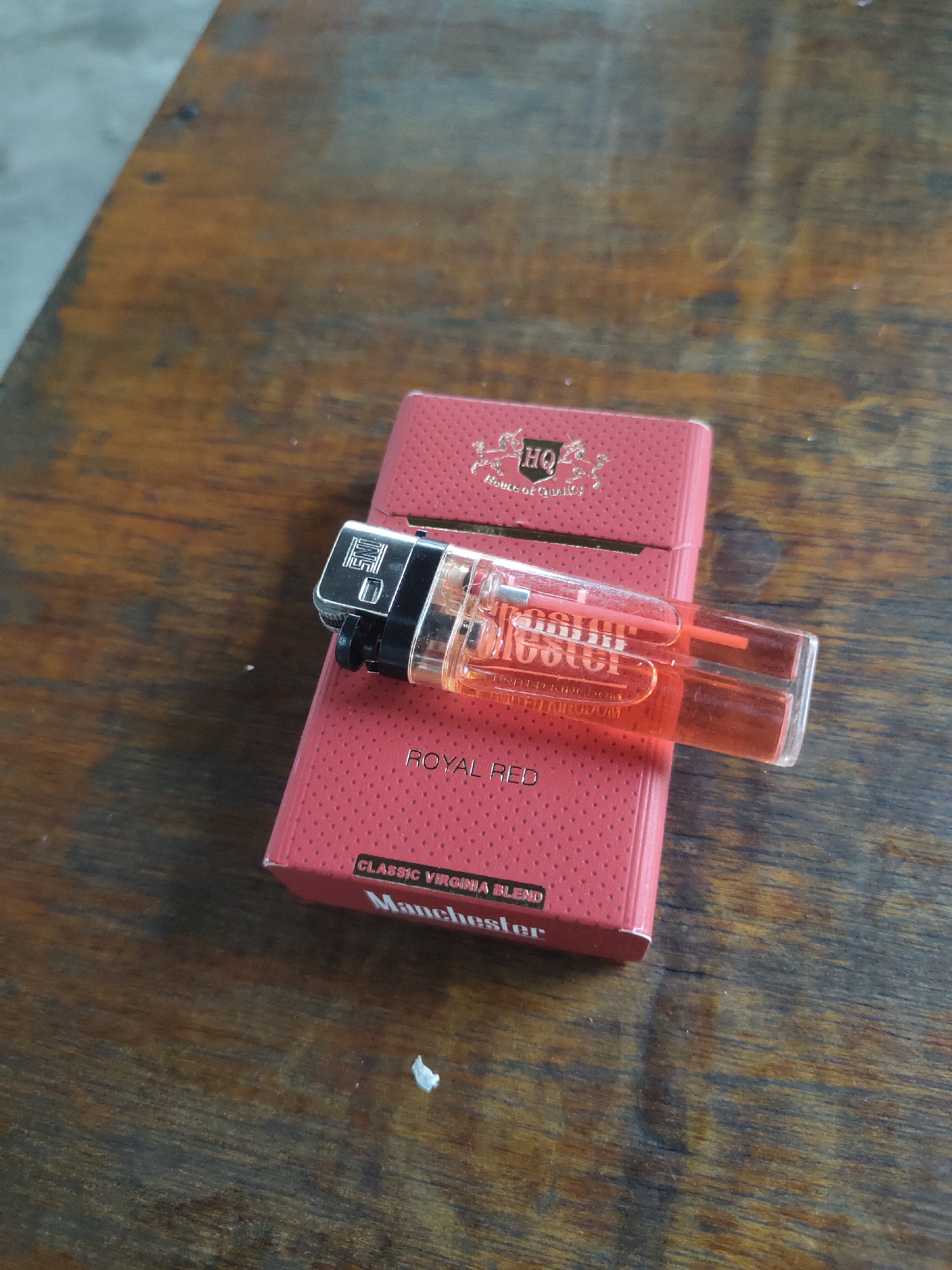 The image displays a red cigarette pack and a clear, orange-tinted lighter resting on a wooden surface. The scene is static, with no activity taking place, suggesting the items are simply laid out.
The cigarette pack is predominantly red with a fine dotted texture. On its front, a gold-colored emblem features the letters "HQ" above the words "House of Quality." Below this, "ROYAL RED" is printed in white, and at the very bottom, "CLASSIC VIRGINIA BLEND" is visible, with the brand name "Manchester" partially shown beneath it.
Positioned diagonally across the cigarette pack is a transparent lighter filled with orange or red fluid, indicating it's a gas lighter. The lighter has a black top and a silver-colored metal cap on its igniter mechanism, which features an "SM" logo. Text is embossed on the lighter's side, clearly reading "King Master" and "UNITED KINGDOM" underneath, confirming its brand and origin.
The wooden surface beneath the objects appears to be a table or desk, exhibiting a warm brown tone with visible grain and some minor wear or imperfections, suggesting an aged or well-used piece of furniture. The lighting is even, implying an indoor setting without direct sunlight, and the time of day is not specifically identifiable. The overall impression is a casual still life. This image is set in Keudematangkuli, Indonesia.