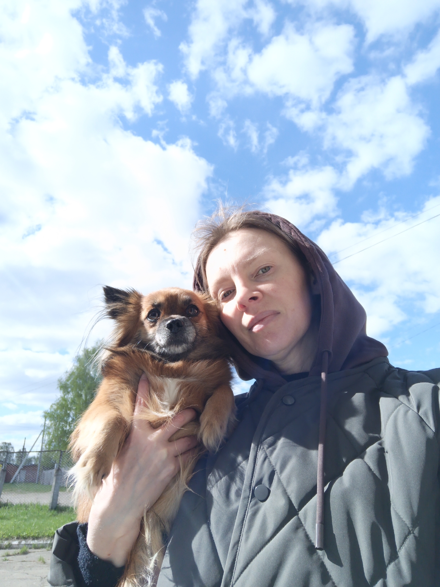 A woman in a quilted jacket stands outside, holding a small, brown dog with long fur. The woman wears a hooded jacket. The jacket is dark green or gray. The sky is blue with puffy white clouds. Grass and a fence are visible in the background. The location is the city of Chaikovsky, Russia.