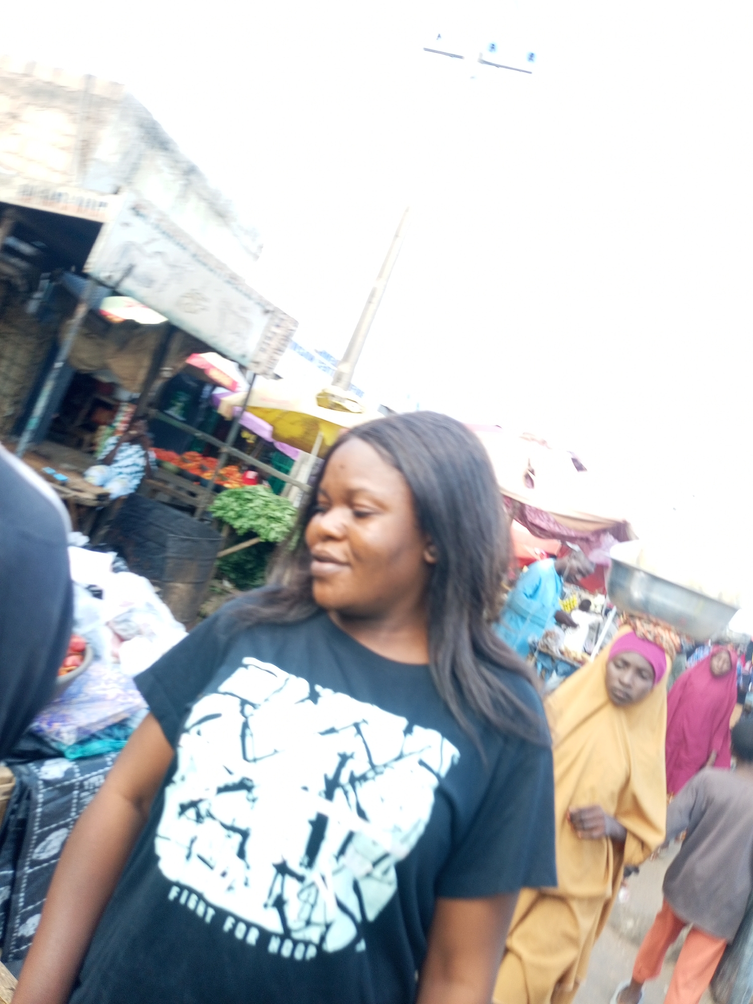 This is a photograph taken at a marketplace in Tempure, Nigeria. In the foreground, a young woman is looking to her left. She is wearing a black t-shirt with a white graphic design and the text "FIGHT FOR HOOP" printed on it. Her hair is dark and falls past her shoulders. To the left of the main subject, partially visible, is another person wearing a black shirt. In the background, several other people are seen. One person, wearing a bright yellow hijab and dress, is looking down. Another person, wearing a pink hijab and dress, is visible behind them. A child in a grey shirt and orange pants is walking away from the camera. The marketplace appears to be outdoors, with stalls and awnings visible. Produce, such as green leafy vegetables and what appear to be tomatoes, are displayed at one of the stalls. The environment is bright, suggesting daylight.