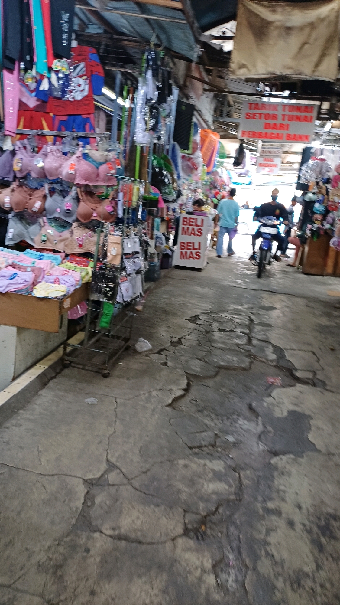 The image captures a scene within an open-air market in Citeureup, Indonesia, characterized by its weathered concrete ground and overhead coverings. Various stalls are visible, displaying a range of goods including undergarments, children's clothing featuring cartoon characters, and other assorted items. A prominent sign with red lettering states "BELI MAS" (Buy Gold) in duplicate, indicating a commercial activity. In the mid-ground, two individuals are observed; one is standing and facing away from the camera, while another is seated on a motorcycle, also facing away. The environment appears to be a bustling marketplace with a mix of goods, signage, and people engaged in daily activities. The ground surface shows significant cracking and wear, suggesting an established and possibly aged infrastructure.