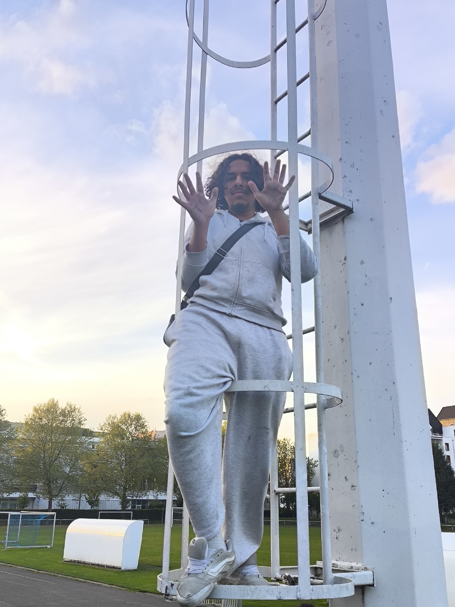 A person is climbing a white metal ladder attached to a tall, square post, likely part of a lighting structure or similar apparatus. The individual is wearing a light grey tracksuit and light-colored sneakers, and has a dark shoulder bag. They are holding their hands up with spread fingers, possibly interacting with the ladder structure. The background shows a sports field with goalposts, a white dugout, and running track. Trees are visible in the middle distance with buildings behind them. The sky is partly cloudy with a mix of blue and pale yellow. The photograph was taken in the city of Schiltigheim, France.