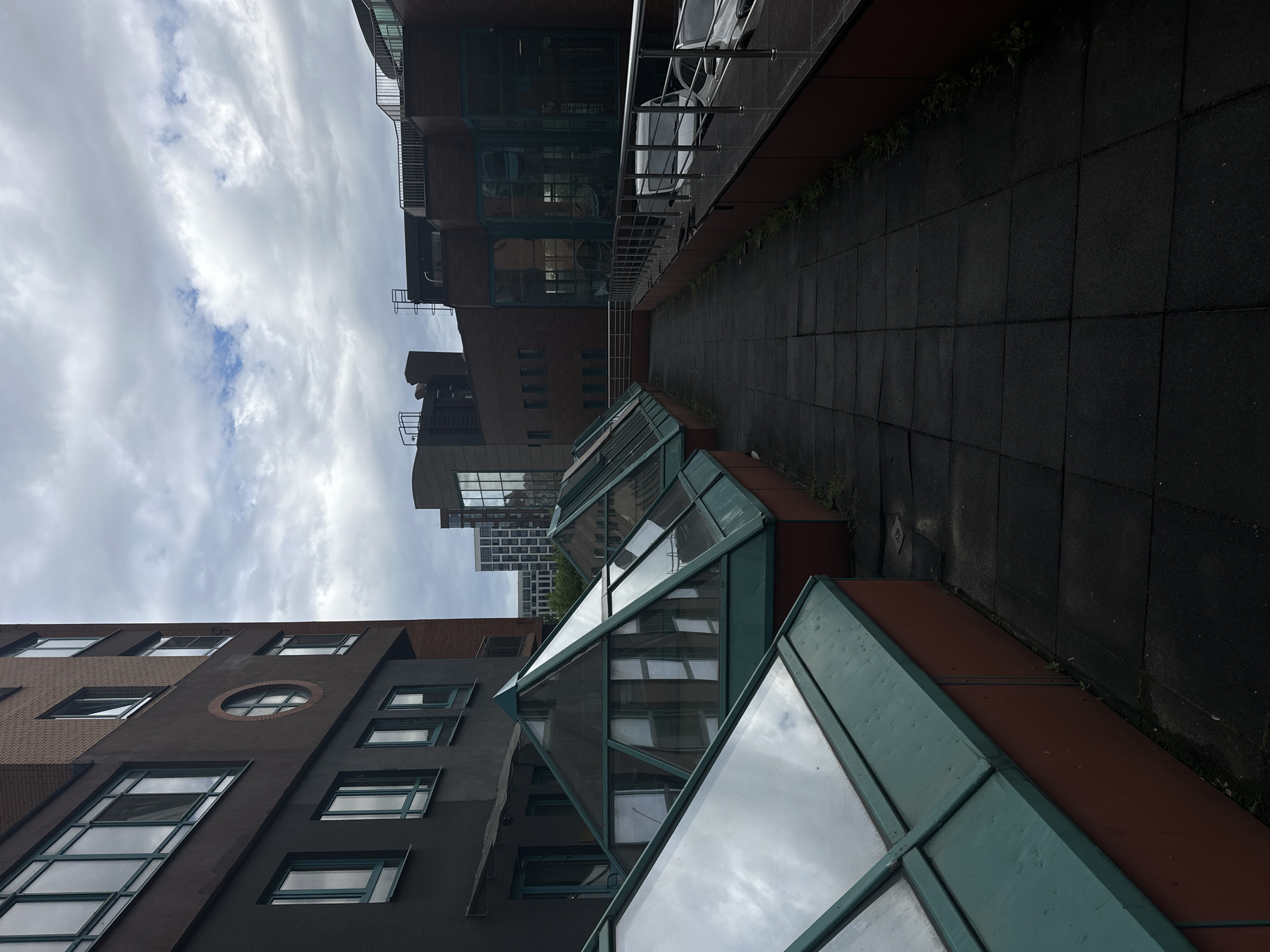 This is a photograph of a modern architectural building with a dark facade and angular, glass-paneled extensions on the lower levels. The building appears to be made of dark brick and concrete, with a mix of rectangular and circular windows. The glass structures have a teal colored frame and reflect the cloudy sky. A dark, tiled walkway runs alongside the building. In the background, another larger building with many windows is visible. The sky is overcast with white and gray clouds, suggesting it may be daytime, possibly morning or afternoon. There are no people or distinct objects visible, focusing solely on the architecture and the environment. The setting appears to be urban, given the presence of multiple buildings and a walkway. There is no visible text in the image. The overall mood is somewhat subdued due to the cloudy weather and the dark tones of the building.