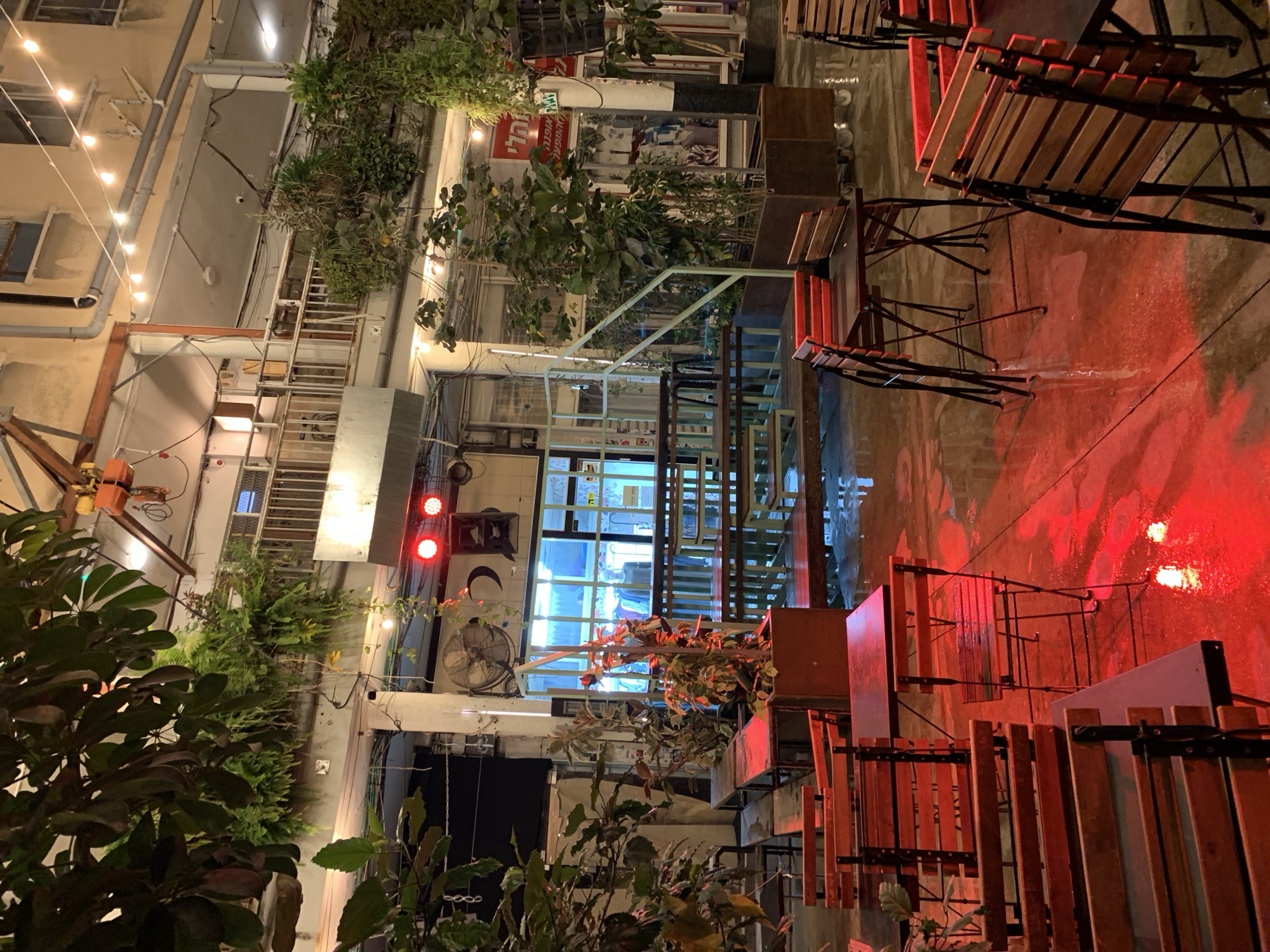 The image depicts a narrow alleyway in Tel Aviv, Israel, at night. The alley is illuminated by overhead string lights and red spotlights, which cast a red hue on the wet pavement. Tables and chairs, primarily made of wood and metal, are arranged on the right side of the alley. Plants and greenery line the walls and hang from above, adding a natural element to the urban setting. The buildings surrounding the alley are varied in style, with visible windows, balconies, and architectural details. A lit storefront or restaurant interior is visible further down the alley, suggesting a bustling atmosphere.