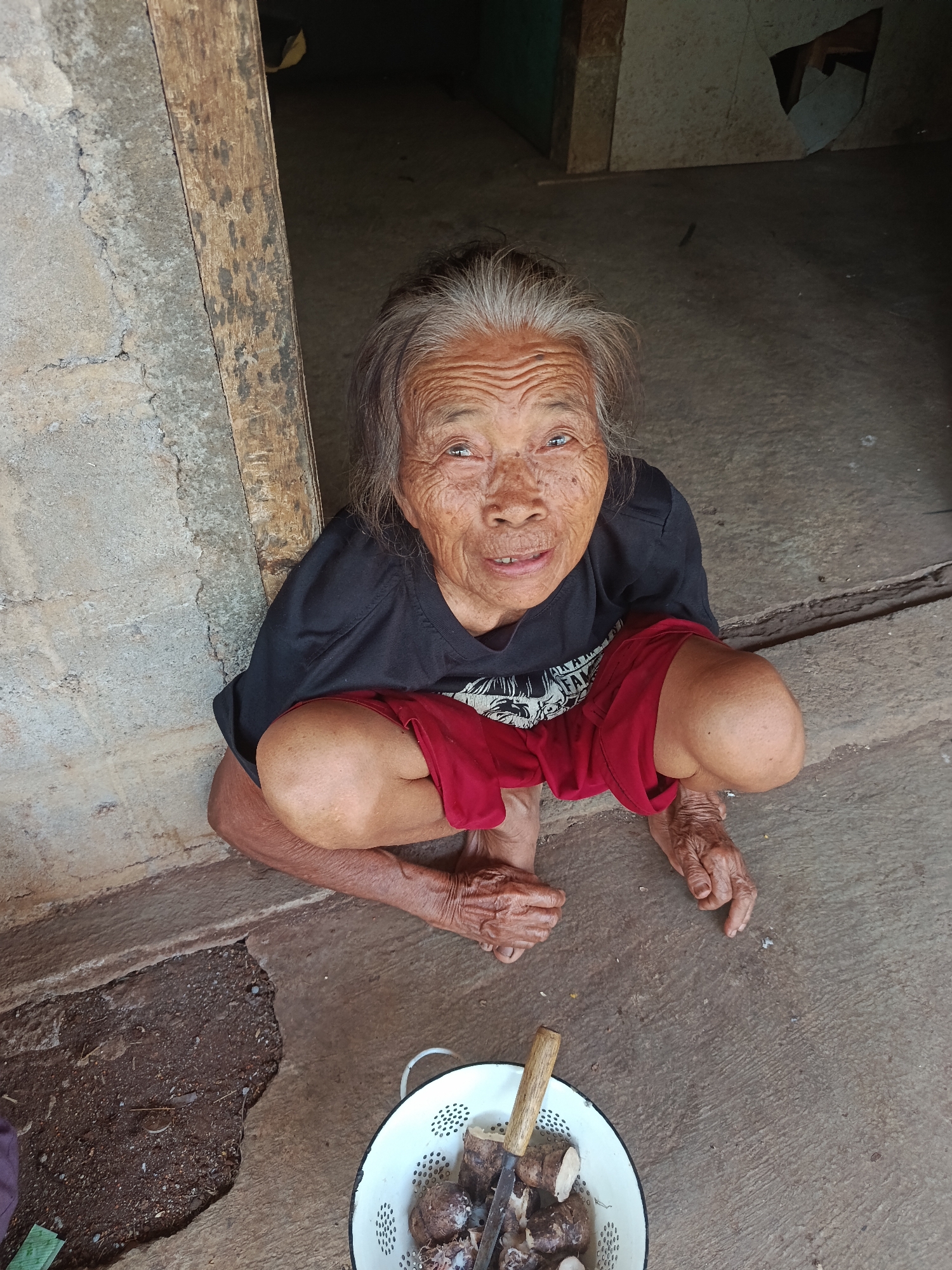 In Karangmojo, Indonesia, an elderly Indonesian woman with wrinkled skin and graying hair crouches on a concrete floor. She is wearing a black t-shirt and red shorts. Her hands are resting on her knees. In front of her, to the right, is a white colander filled with root vegetables and a knife. The background appears to be the entrance to a dwelling, with rough concrete walls and a wooden doorframe.