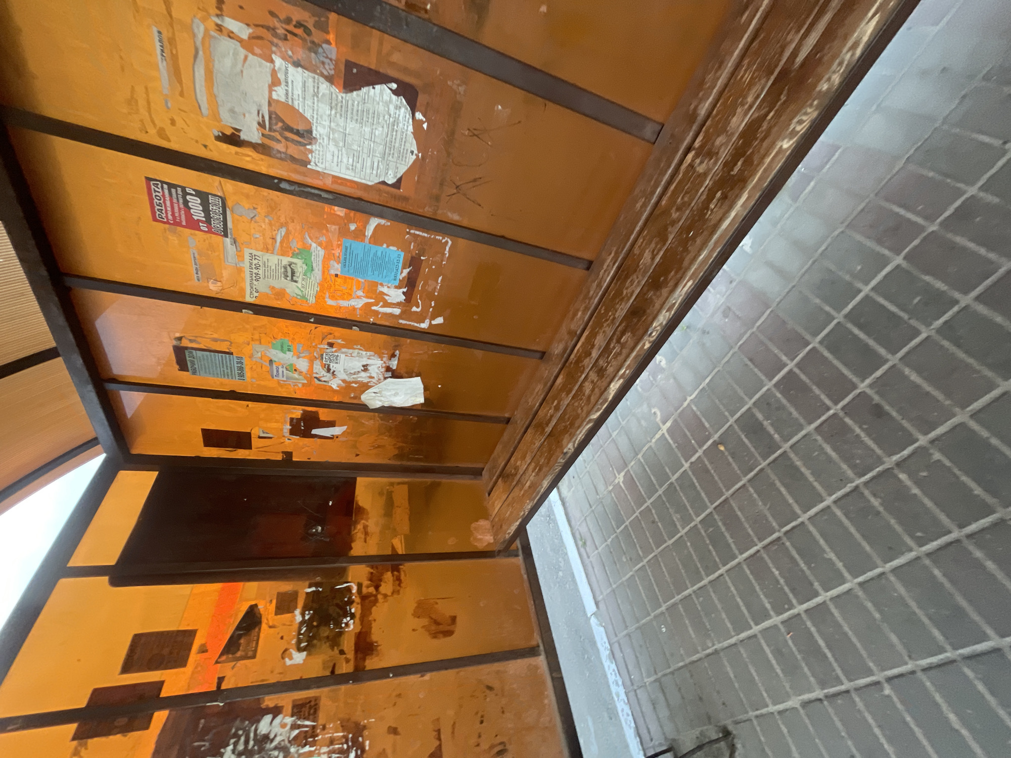 The image shows an interior view of an orange bus stop shelter with a wooden bench. The walls of the shelter are made of orange translucent panels, some of which have advertisements pasted on them. The floor is paved with gray square tiles. The shelter is located in the city of Krasnye Baki, Russia. The scene is deserted.