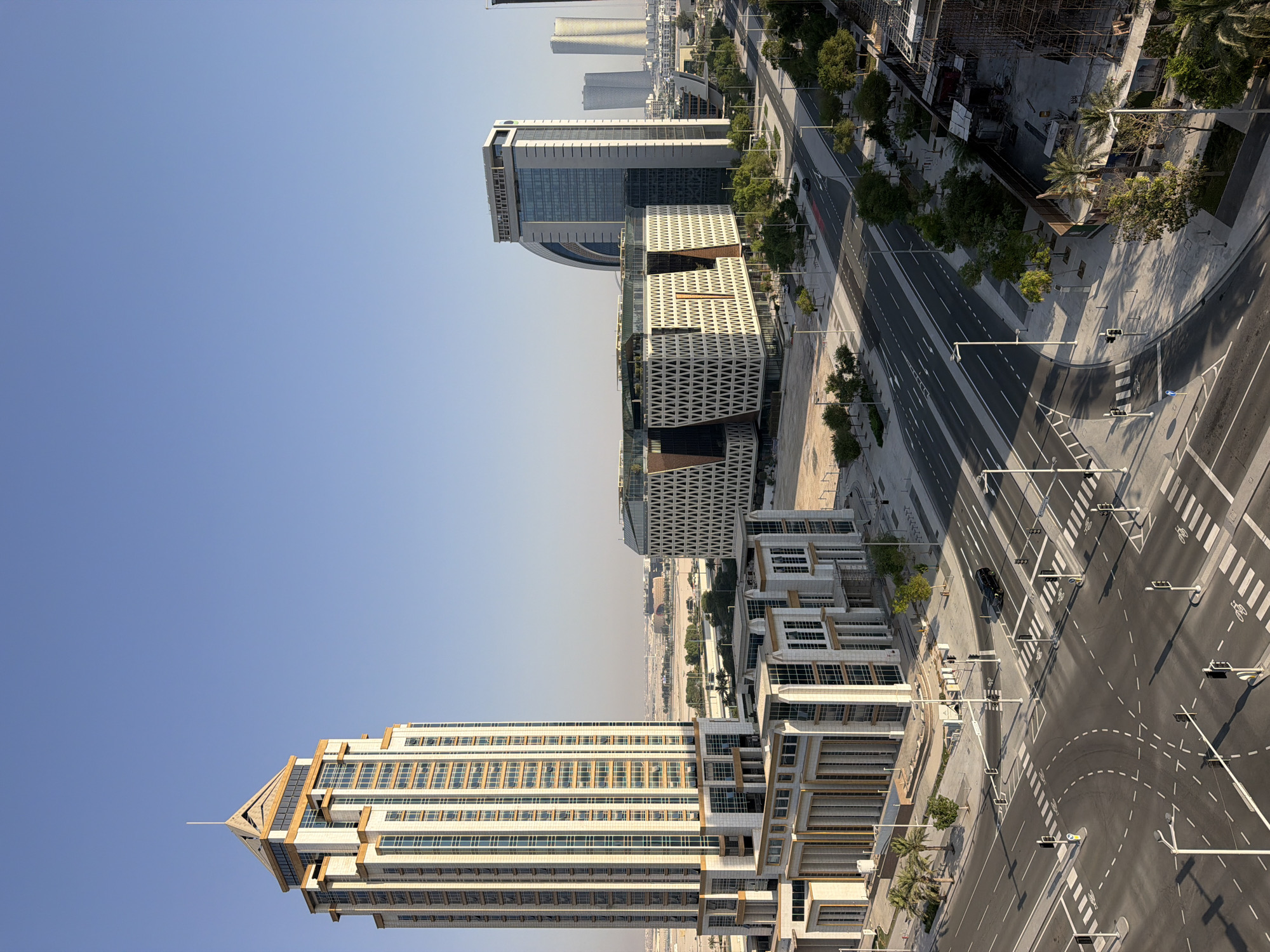 A high-angle, full shot shows a modern city intersection with several large, contemporary buildings under a clear, pale blue sky. In the foreground, a complex intersection with multiple lanes and crosswalks dominates the scene. Traffic signals and streetlights are visible, with a single black SUV navigating the intersection. Sidewalks lined with sparse trees and palm trees border the roads.
To the left, a tall, ornate skyscraper with a tiered, pointed roof stands prominently. Its facade features a grid of windows with golden frames, set against a light-colored stone or concrete exterior. In front of this skyscraper is a lower, rectangular building with a classical design, featuring columns and large windows.
To the right of the intersection, a uniquely designed building with a complex geometric facade made of white lattice-like structures and dark accents is visible. Behind this, a taller, sleek skyscraper with a curved, glass facade rises, and further in the distance, two very tall, slender, curved skyscrapers are visible. The overall environment suggests a sunny day in a developed urban area with ongoing construction in some parts.