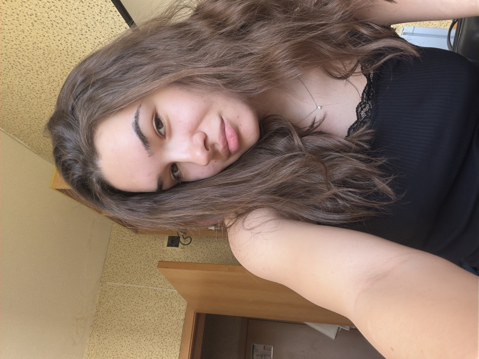 A young woman with long, wavy, medium-brown hair is captured in a close-up selfie, taken indoors in Sofia, Bulgaria. She is lying down, resting her head on her right arm, and looks towards the camera with a gentle, relaxed expression and a soft, faint smile. Her brown eyes are open, and her lips are slightly parted. She is wearing a black ribbed tank top with a delicate lace trim along the neckline. Around her neck, a thin silver chain necklace with a small, clear stone pendant is visible.
Her abundant hair, parted on the side, frames her face and flows around her shoulders. The background suggests an indoor setting with varied textures. To her left, a plain, light beige wall is visible. Behind her, both the wall and ceiling feature a consistent light yellow or beige speckled texture. In the lower right of the frame, a closed wooden door with a light brown finish is prominent. On the speckled wall near the door, a small, white rectangular object, possibly a sign or sticker, is visible, though any text on it is unreadable. Above the door, a dark electrical fixture or outlet can be seen. The lighting is soft and appears to be natural daylight. The overall impression is calm and introspective.