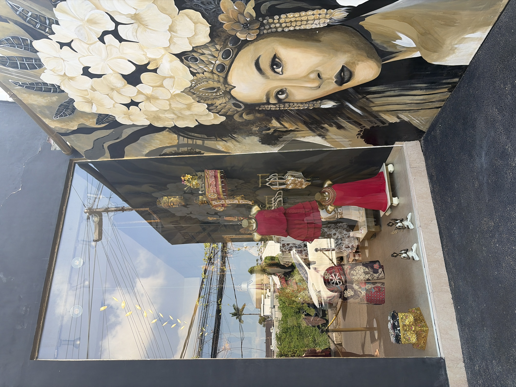 The image shows a storefront window display in Kuta, Indonesia. The display features mannequins dressed in red and patterned clothing, as well as decorative boxes and small statues of figures in meditation poses. A large mural of a woman wearing a headdress is painted on the exterior wall to the right of the window. Reflections in the window show the sky, overhead wires, and distant buildings and palm trees. A white car and a scooter are visible through the window, suggesting the storefront is located on a street. The overall scene conveys a sense of an eclectic retail environment, possibly selling clothing or artisanal goods.