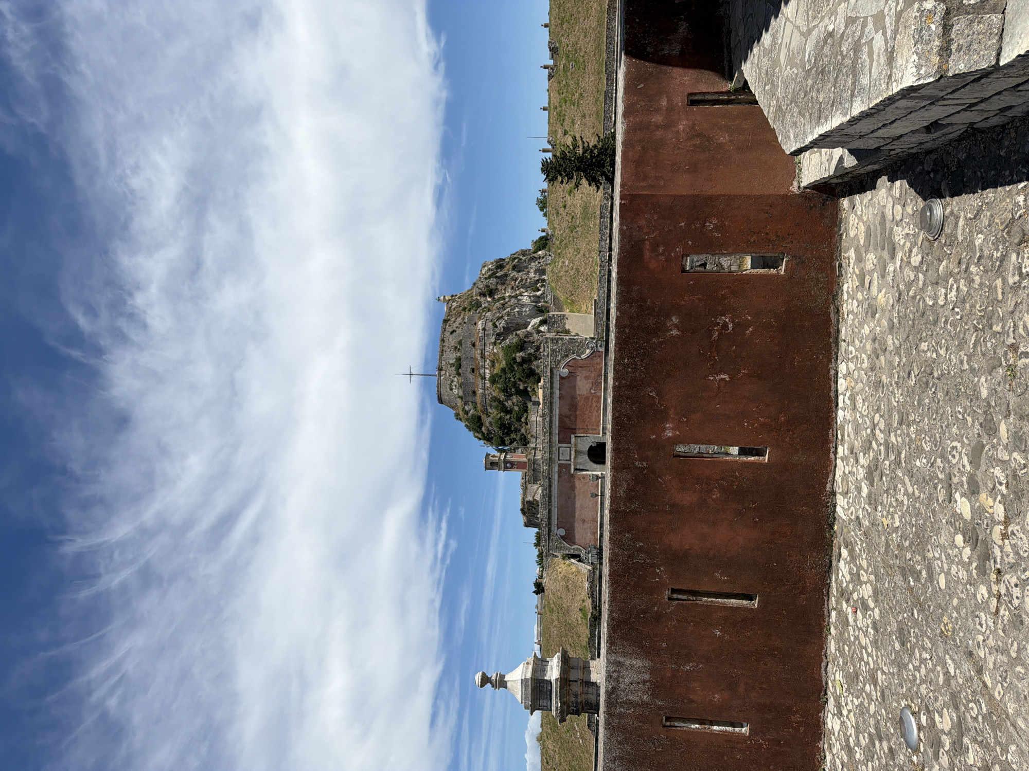 The vertically oriented image presents a historical fortified landscape under a bright, partly cloudy sky. In the immediate foreground, a weathered reddish-brown stone or concrete wall extends across the frame, featuring several regularly spaced, narrow, rectangular openings, likely embrasures or small windows. To the right of this wall, a textured stone pathway or ground, composed of various light-colored pebbles, is visible, with sunlit and shadowed areas. Several small, circular, metallic ground lights are embedded in this pavement.
Beyond the foreground wall, a grassy, gently sloping terrain leads to more substantial stone structures of a fortress. In the midground, parts of the fortification complex are visible, including extensive stone walls, buildings, and a distinctive clock tower with a red roof and white clock faces. A decorative, ornate stone finial or pinnacle is prominently placed on a wall in the lower-left midground. A solitary dark green tree stands on the grassy slope.
The background is dominated by a steep, rugged, rocky hill, which appears to be part of the citadel, crowned with additional fortifications and a prominent cross at its summit. The sky above is a vibrant blue, adorned with sweeping, wispy white cirrus clouds. The scene is illuminated by bright daylight, casting distinct shadows and suggesting either morning or afternoon. No people are present, and no specific activity is occurring. The location strongly resembles the Old Fortress in Corfu, Greece. There is no visible text in the image.