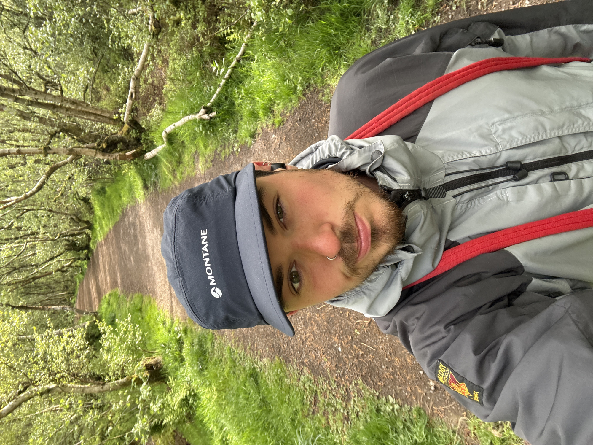 A medium shot depicts a man wearing a Montane cap, a gray and black jacket with red accents, and a nose ring. He is standing on a dirt path lined with green vegetation and trees, which appear to be in a forest or park. The location is identified as Unknown, Unknown.
