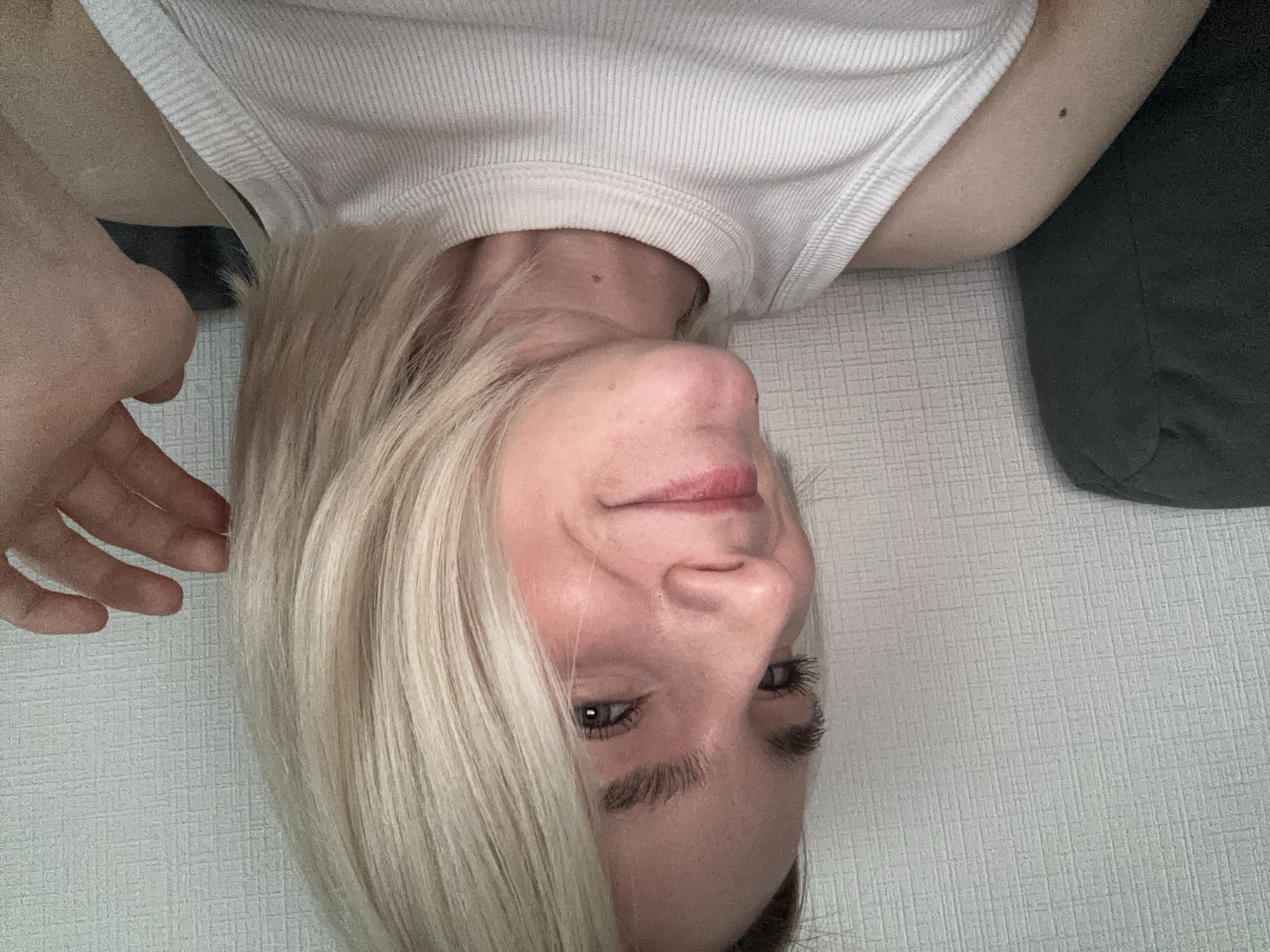 A fair-skinned person with short, platinum blonde hair and light-colored eyes is oriented upside down in the frame. They wear a white tank top. Their left hand is raised to the side of their head. To the right is a dark grey, soft object, likely a cushion or piece of furniture. The background appears to be a neutral-toned, textured surface, possibly a wall or bedding. Location information is unavailable.