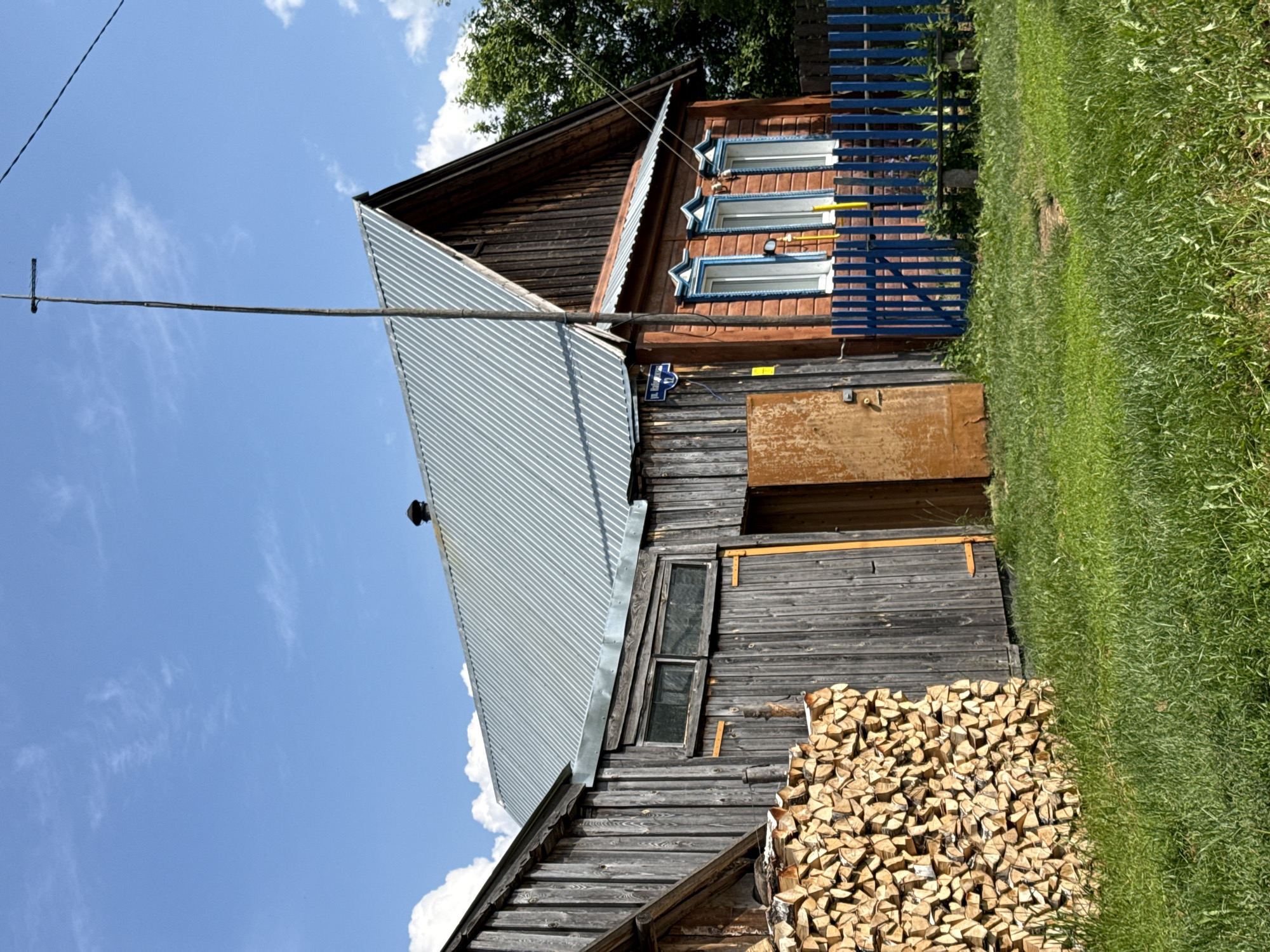 The image captures a sunny day scene in the city of Zasechnyi, Russia, featuring a rural or semi-rural setting with traditional wooden buildings. In the foreground, a neatly stacked pile of chopped firewood is visible next to a weathered wooden structure, likely a shed or barn, with vertical plank siding and a metal roof. To the right, a two-story wooden house painted a warm orange hue stands with three windows adorned with decorative blue trim. A blue picket fence encloses the yard in front of the house. A tall, thin antenna on a wooden pole extends upwards, piercing the bright blue sky dotted with white, fluffy clouds. The ground is covered in lush green grass. The overall impression is one of a simple, well-maintained rural home.