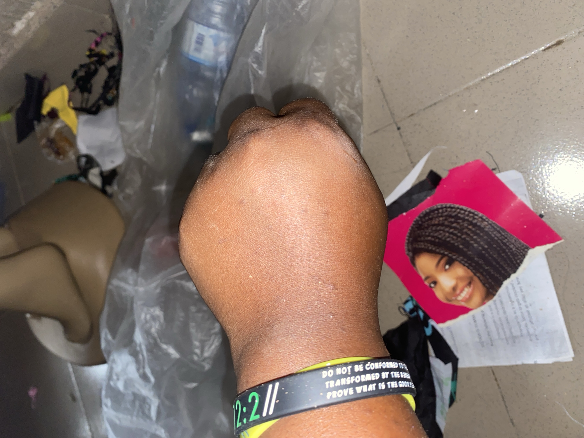 The image depicts a close-up of a person's clenched fist, adorned with a black and green wristband. The wristband has text that is partially legible, appearing to say "DO NOT BE CONFORMED TO THIS WORLD. BE TRANSFORMED BY THE RE… PROVE WHAT IS THE GOOD." Behind the fist, a torn piece of paper with a photograph of a smiling woman with braided hair is visible, placed on a tiled floor. In the background, several plastic bags, a brown mannequin leg or torso, and various other items are scattered, suggesting a cluttered environment. The lighting is dim, and the overall scene appears to be indoors, possibly in a storage area or a discarded pile. The specific location in Jauro Yinu, Nigeria, cannot be determined from the visual information provided.