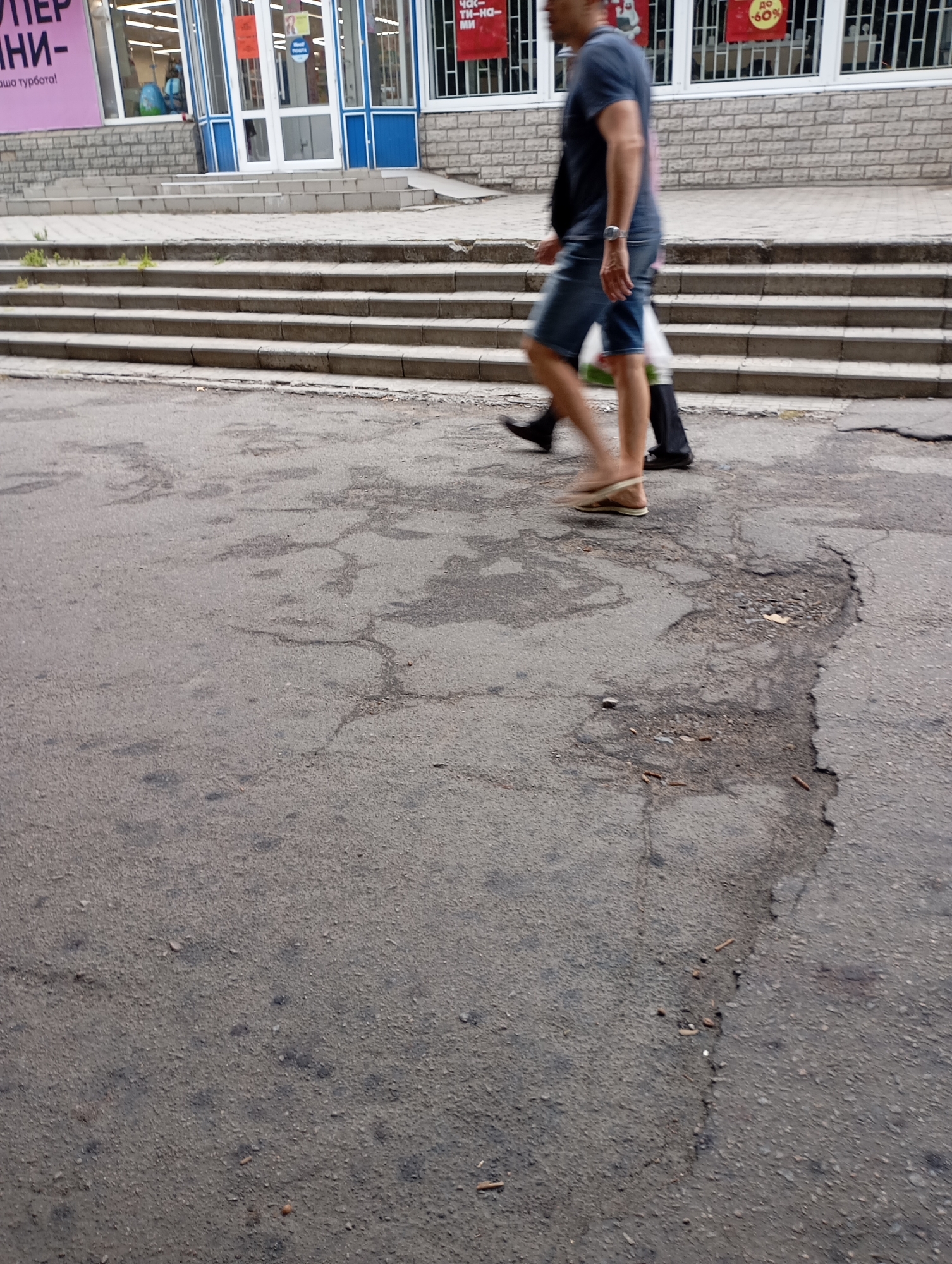 A medium-angle, eye-level shot captures a street scene in Horishni Plavni, Ukraine, with a focus on a cracked asphalt surface in the foreground. Two individuals are captured in motion, walking across the uneven pavement. The person in the foreground, on the left, is wearing denim shorts and a dark t-shirt, with their right leg slightly forward, clad in a flip-flop. They are carrying a white plastic bag in their left hand. Behind them, to the right, another individual is walking, their legs obscured by motion blur, wearing dark pants and dark shoes. In the background, a building with large glass windows and blue trim is visible, with a set of concrete stairs leading up to its entrance. Several signs are present on the building, including a large pink sign on the far left and circular red signs with white text indicating discounts. The environment appears to be an urban commercial area, with evidence of wear and tear on the road surface.