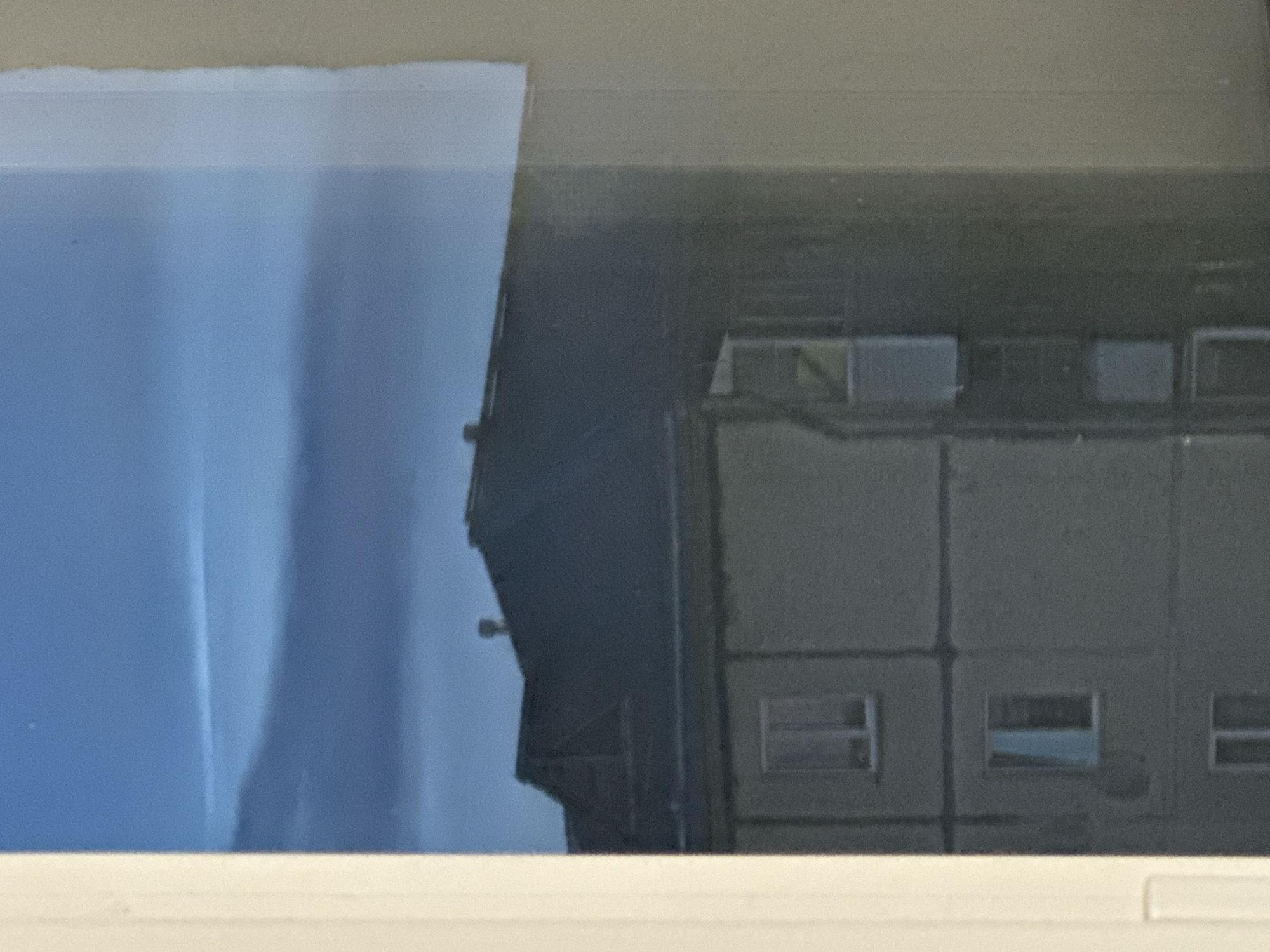 The image captures a view through a window, showing a residential building with a dark-colored roof and a grey facade. Multiple windows are visible on the building's exterior, some with reflections of the sky. The sky itself is a deep blue with faint cloud formations. The scene appears to be in Plesetsk, Russia, with the building likely being a multi-story apartment block. The left edge of the image is framed by a light-colored window frame. No people or overt actions are depicted, creating a still and observational scene.