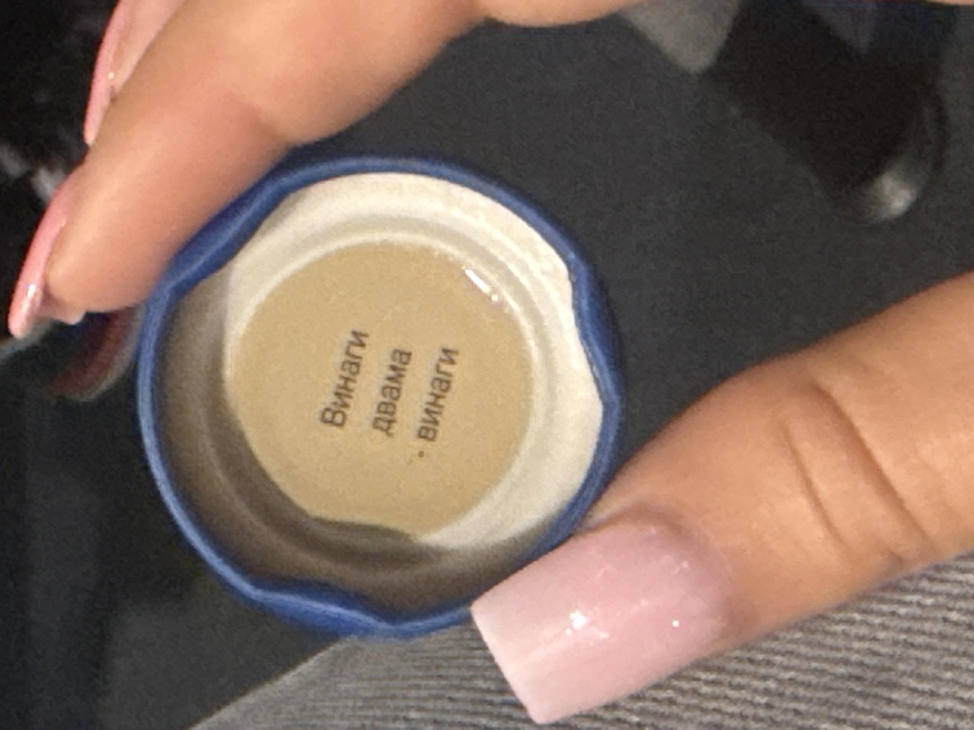 A close-up, over-the-shoulder shot shows a person's hands holding a blue plastic bottle cap with text printed on a light brown insert. The text reads "Винаги двама - винаги" which translates from Bulgarian to "Always two - always". One hand, with light pink, squared-off nails, is visible in the lower left, holding the cap open. The other hand, with slightly longer, shiny pink nails, is partially obscuring the upper right portion of the cap. The background is dark and out of focus, suggesting an indoor setting. No specific location in Rehestädt, Germany, can be identified from the image.