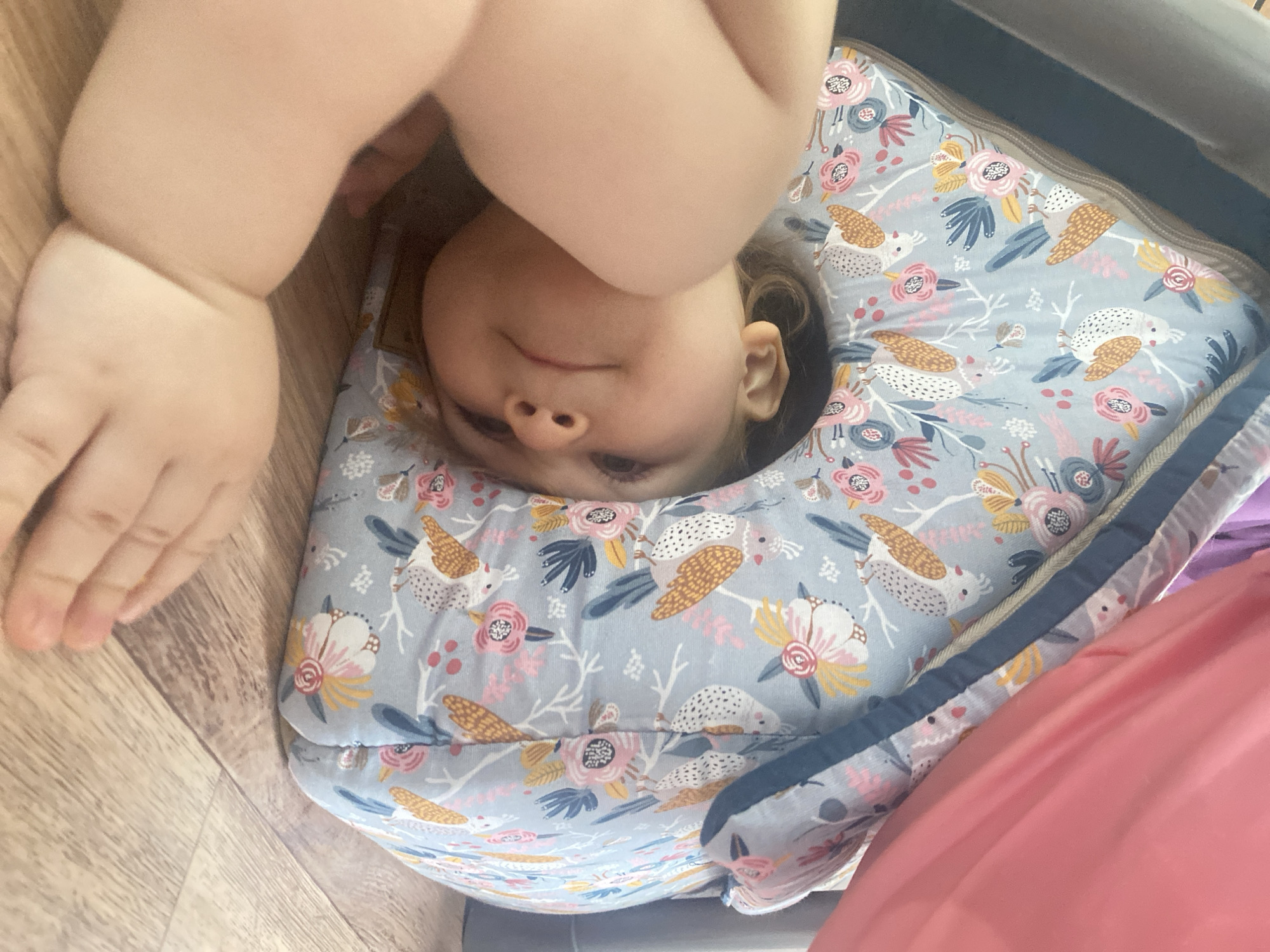 A child is upside down, with their head encircled by a patterned, soft, ring-shaped object. The child's face is visible, with eyes open. Part of an arm and hand are visible at the top left, with the fingers slightly curled. The ring-shaped object is patterned with images of flowers and birds on a light blue background. A wooden floor is visible in the lower left corner, and a pink object is in the lower right corner. The location is unknown.
