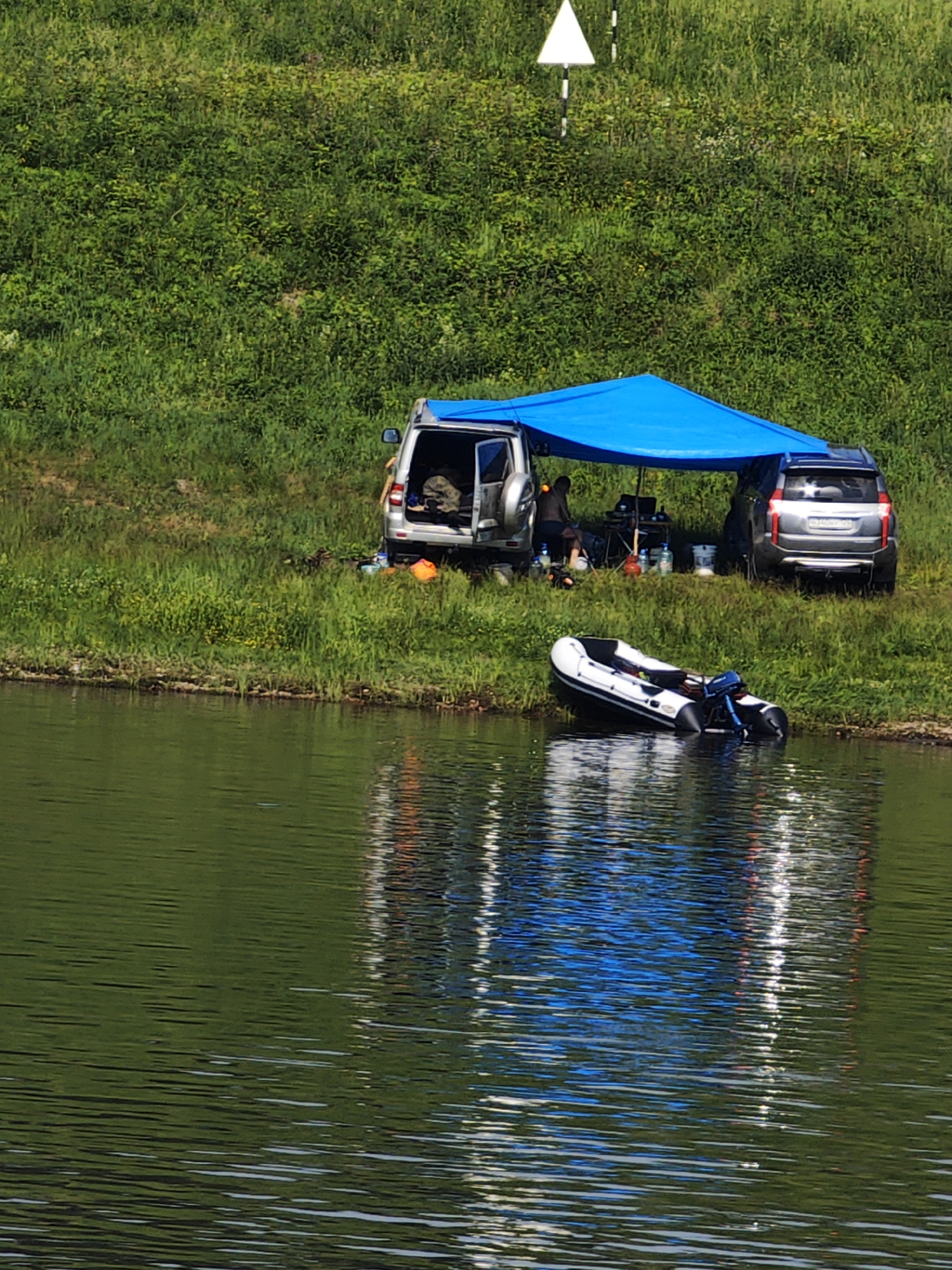 The image depicts a daytime scene near a body of water, likely a river, in Podporozh'e, Russia. The foreground is dominated by the greenish water with ripples, reflecting the environment. Along the bank, there is a grassy area with some vegetation.
On the grassy bank, two silver SUVs are parked next to each other. A bright blue tarp is stretched over them, creating a shaded area. Under the tarp, there appears to be some camping or fishing equipment, including what looks like a small table and chairs. A person is visible sitting under the tarp, facing away from the camera. Several bottles and containers are scattered in the vicinity.
In the water, near the bank, an inflatable boat with a motor is partially submerged. The boat is white with black accents.
In the background, a steep, densely vegetated grassy hillside rises from the bank. There is a white triangular marker with a black pole, possibly a navigational aid or marker, visible near the top of the hill. The overall impression is a scene of outdoor recreation by the water.