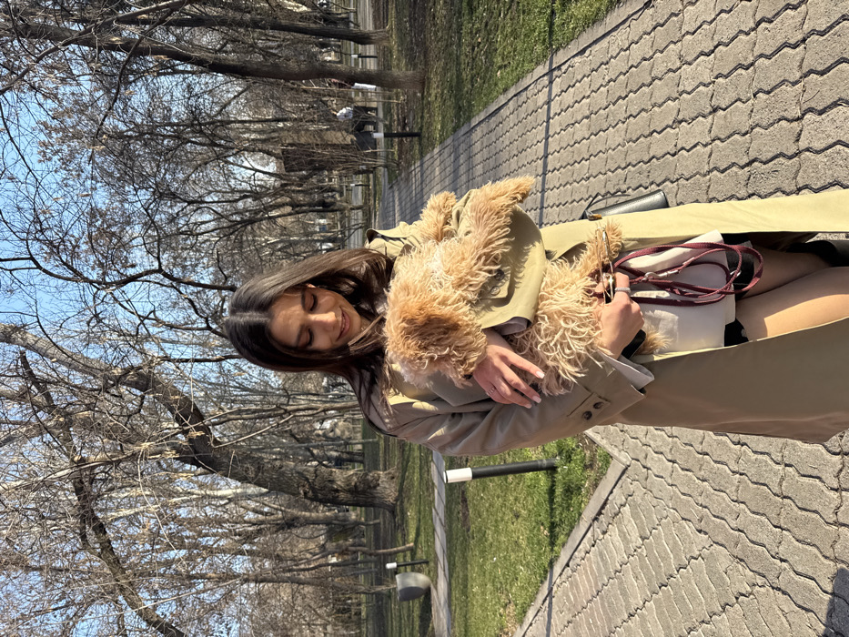 The scene depicts a female subject in a park in Yerevan, Armenia. The subject, with long dark hair, is positioned centrally on a paved pathway, looking downwards with a slight smile. She is wearing a light beige trench coat over a white top and dark shorts or a skirt, with dark tights or stockings and dark footwear. A silver-colored ring is visible on her left ring finger. She holds a small, light brown, curly-haired dog in her arms. A dark red leash is present, draped from the dog or held in her hand, along with a small dark shoulder bag hanging on her right side.
The park environment features a paved pathway constructed from interlocking grey and beige rectangular pavers. Green grass areas are visible bordering the path. Numerous deciduous trees with bare branches dominate the background, indicating a leafless season. The sky above is clear blue. Several modern, dark-colored park lights are spaced along the pathway and in the grassy areas. In the deeper background, multiple indistinct human figures are visible among the trees, suggesting other park visitors. Bright, natural sunlight illuminates the scene, casting shadows.
