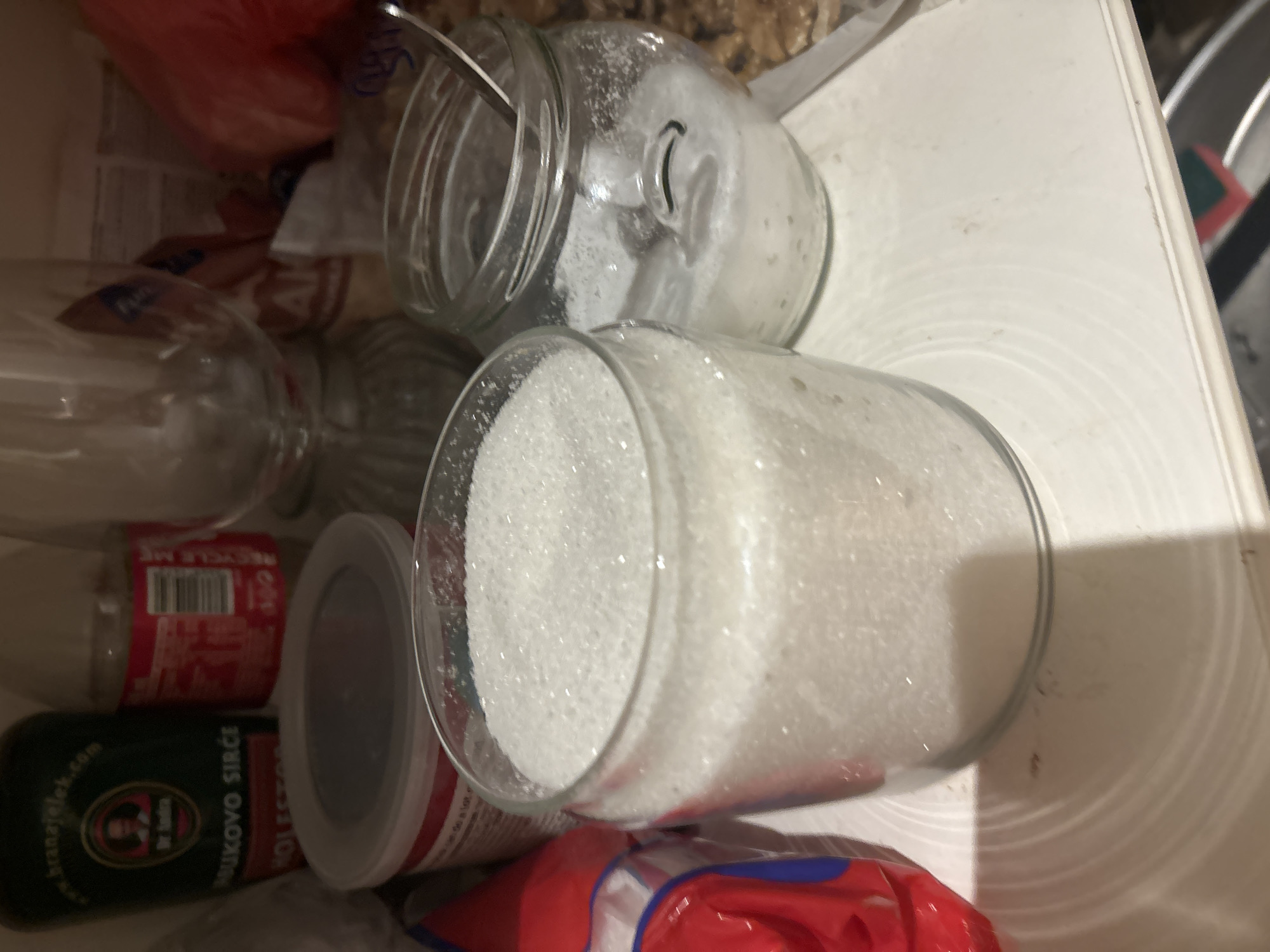 The image depicts the interior of a shelf, likely in a kitchen or pantry, in Budva, Montenegro. The foreground is dominated by two glass jars filled with granular white sugar. The jar on the left is larger and more filled, while the one on the right is smaller and contains less sugar, with a spoon partially submerged in it. To the left of the sugar jars is a red and white cylindrical container with a grey lid, possibly holding more sugar or another ingredient. Behind these, a green bottle with a label in a foreign language is visible, along with a clear glass with a ribbed base, likely an inverted glass. In the background, various packaged goods and containers are stacked, including a red plastic bag on the far left, and what appears to be nuts in a plastic bag on the far right. The overall lighting is dim, suggesting an indoor environment with artificial light. No people are present in the image.