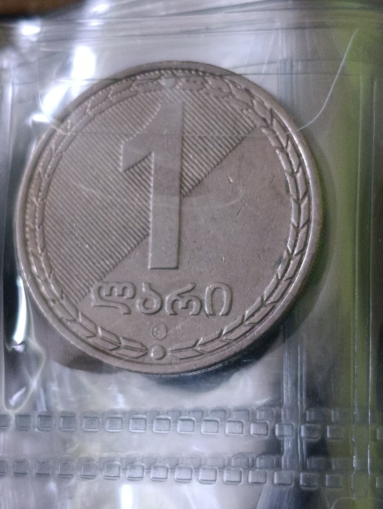 A close-up, top-down shot captures a portion of a coin, encased in a clear plastic sleeve. The coin features a large numeral '1' prominently displayed in the center, with diagonal lines filling the left portion of the circle. To the right of the numeral, Georgian script is visible, along with a laurel wreath encircling the outer edge of the coin. The plastic sleeve exhibits a patterned texture with rectangular indentations. The background is blurred, with hints of green and white light reflecting on the plastic. No specific location in the city of Unknown, Unknown can be determined from the image.