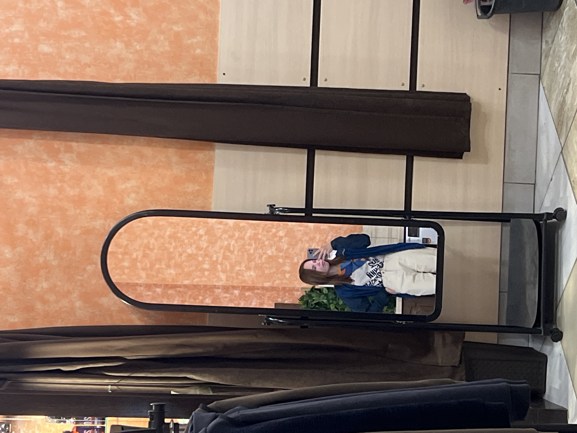 A full-length mirror is centered in the frame, reflecting a woman taking a selfie. She is wearing a blue jacket over a white graphic t-shirt and light-colored pants. A green leafy plant is visible in the background of the reflection. The mirror is framed by a dark metal stand with wheels. In the foreground and behind the mirror, brown and dark blue fabric items are draped on racks, likely clothing. The background wall consists of textured orange paint on the left and light beige panels on the right. Tile flooring is visible to the far right. The location is Verkh-Sosnova, Russia.