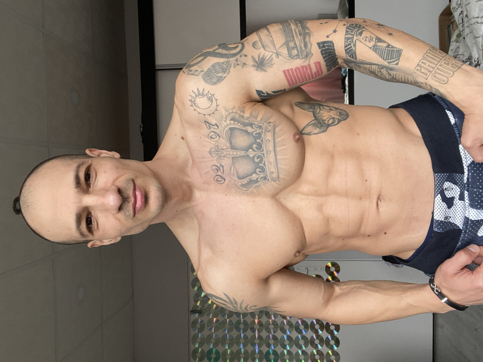 A shirtless man with a shaved head stands indoors, displaying a muscular physique and numerous tattoos on his arms and chest. The tattoos include a crown, the word "WORLD", and other symbols. He is wearing a dark blue and white patterned garment around his waist. A black bracelet adorns his right wrist. The background features a wall, possibly in a residence, decorated with a pattern of circles that could be CDs or similar reflective discs. There is no specific information within the image to determine if the location is Pleven, Bulgaria.