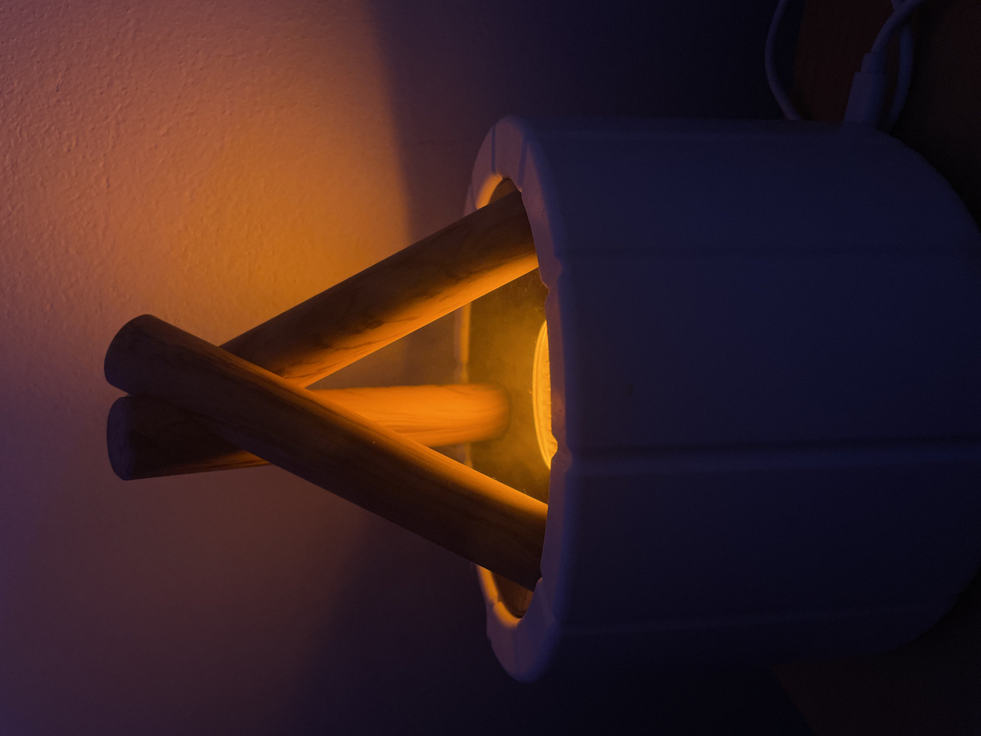 A dimly lit photograph shows a modern wall sconce featuring a cylindrical white lampshade and wooden dowels arranged in an X-shape across the front. An orange-yellow light emanates from within the shade, illuminating the wall behind it. Electrical cords are visible at the top of the fixture. The location is not discernible from the image.
