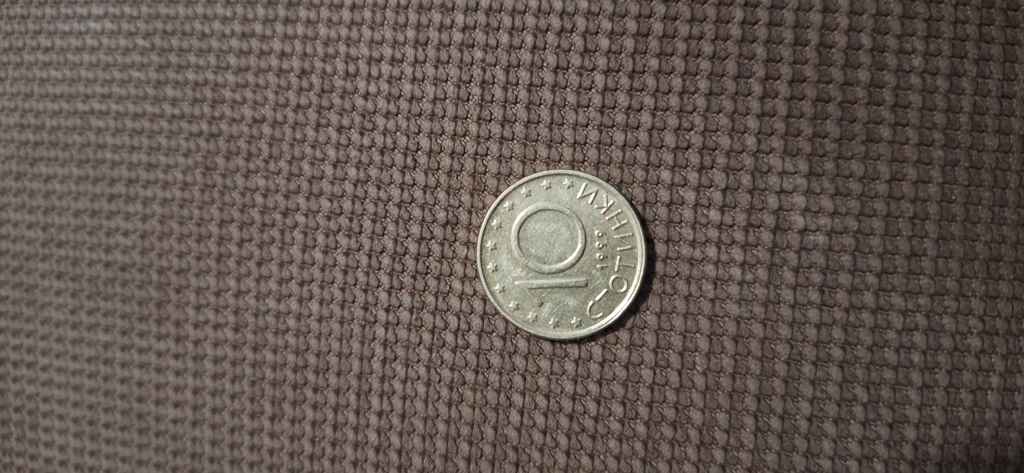 A Bulgarian 10-stotinki coin is placed on a textured, dark brown surface. The coin displays the number "10" and the inscription "СТОТИНКИ" (stotinki), indicating its value. The surrounding text and stars mark it as a Bulgarian currency. No humans or other objects are present in the scene. The location is not identifiable from the image and cannot be reliably specified as Blagoevgrad, Bulgaria. The environment consists solely of the textured surface beneath the coin.
