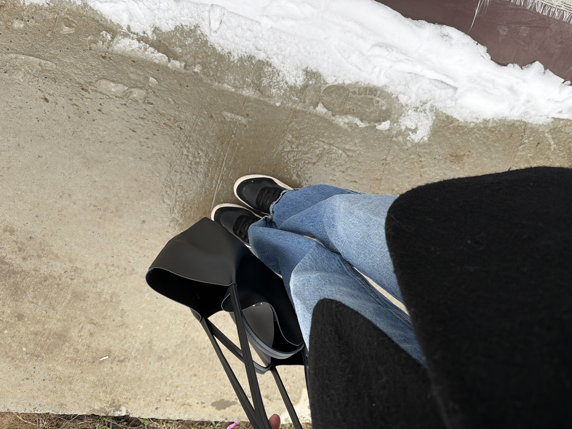 The image shows a person standing on a sidewalk in an outdoor environment. The person is wearing blue jeans and black sneakers with white soles. A black tote bag is positioned next to their legs. The ground is concrete and appears to be wet, with patches of ice and snow along the edge. There is a footprint visible in the snow. The environment is suggestive of winter conditions. The exact location in the city of Chaikovsky, Russia is undeterminable.