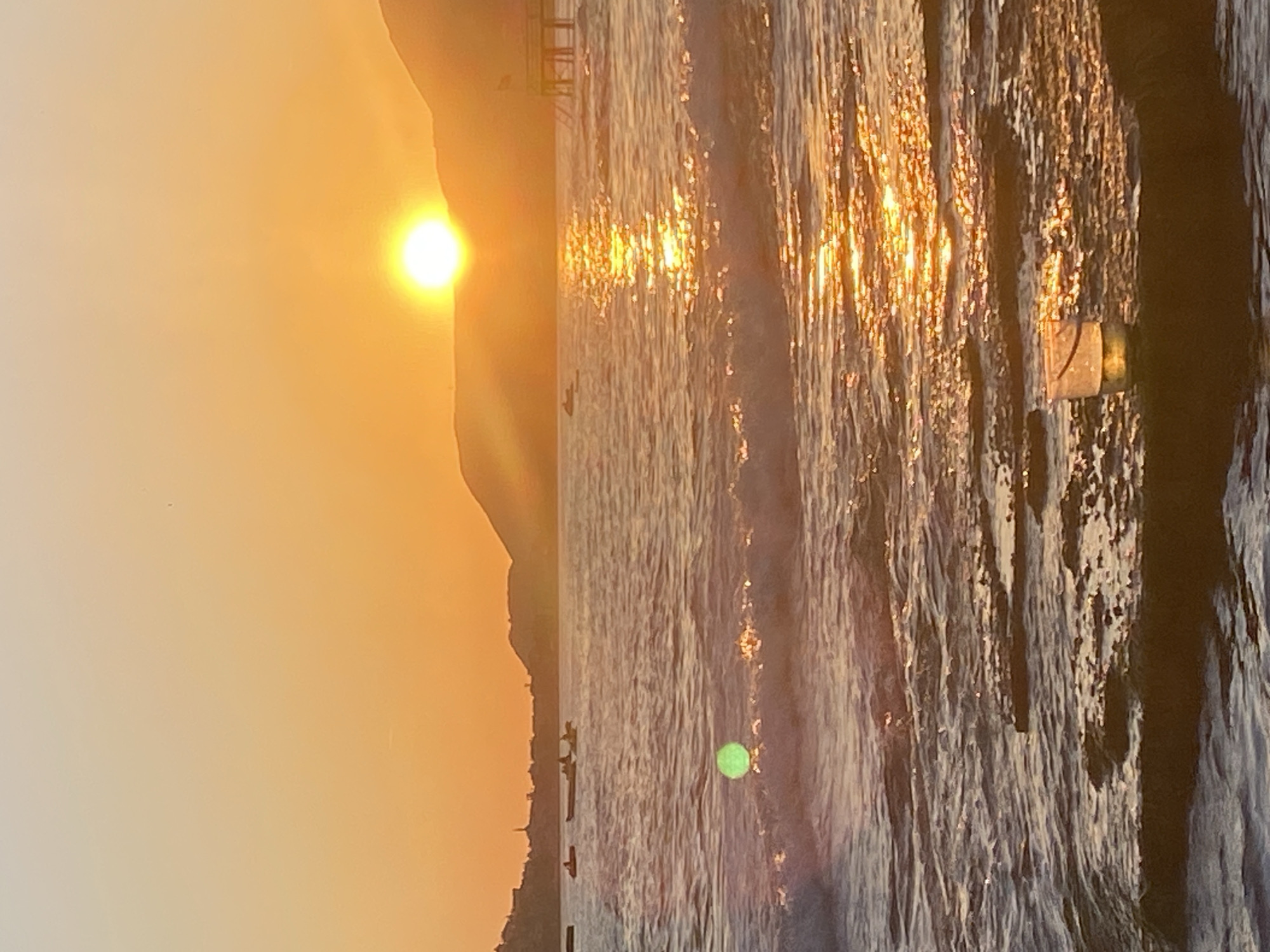 The image captures a sunset scene in Alanya, Turkey. A bright sun is positioned near the top of a mountain silhouette on the left side of the frame. The sea, reflecting the golden light of the setting sun, dominates the right side of the image. The water appears textured with the movement of waves. Some wooden structures or poles are visible along the shoreline on the right, with a cylindrical object attached to one. There are faint silhouettes of buildings or structures along the base of the mountain on the left. The overall tone of the image is warm, bathed in the hues of the setting sun.