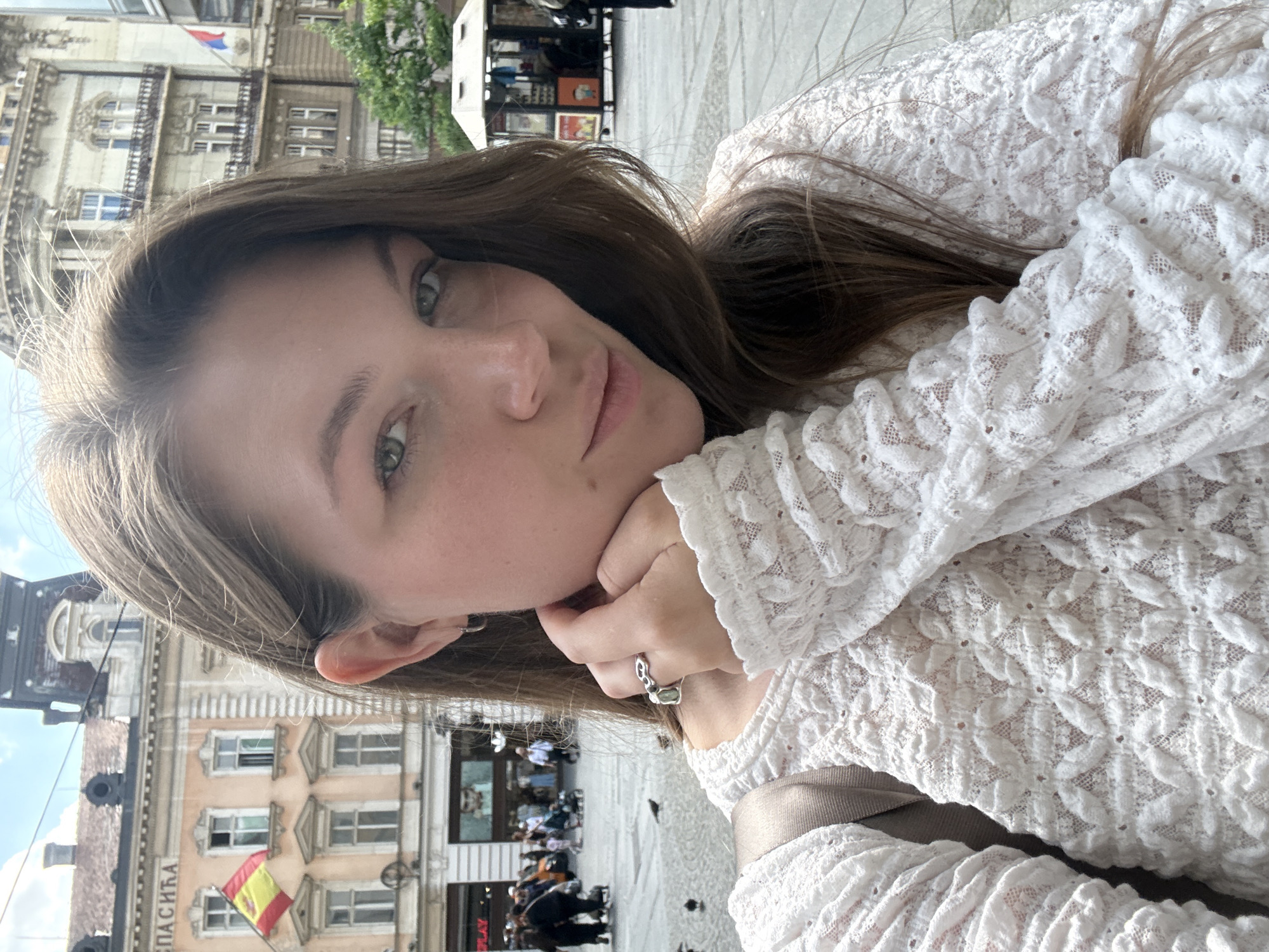 A young woman with brown hair and green eyes is posing for a selfie in Belgrade, Serbia. She is wearing a white lace top and resting her chin on her hand, which has a ring on the finger. In the background, there is an old building with a Spanish flag hanging from it. People can be seen walking around in the background, and there is also a food stall.