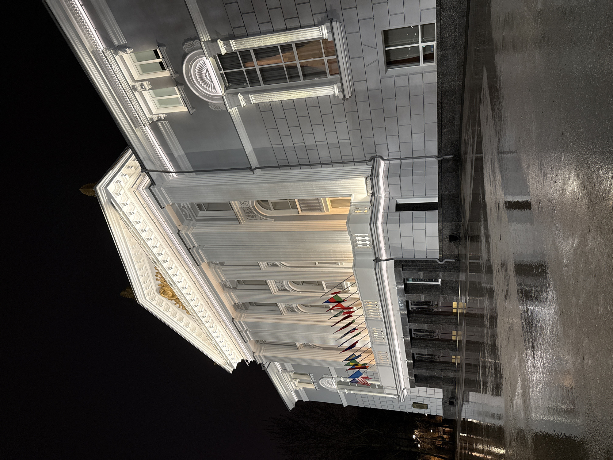 A nighttime photograph captures a building with classical architectural elements, likely located in Perm, Russia. The building is white with prominent columns and decorative trim under the roof. Flags representing multiple countries are displayed along the building's facade. The sky above is dark, suggesting it is night. In the foreground, the ground appears wet and reflective, possibly from rain. The building's reflection is visible on the wet surface.