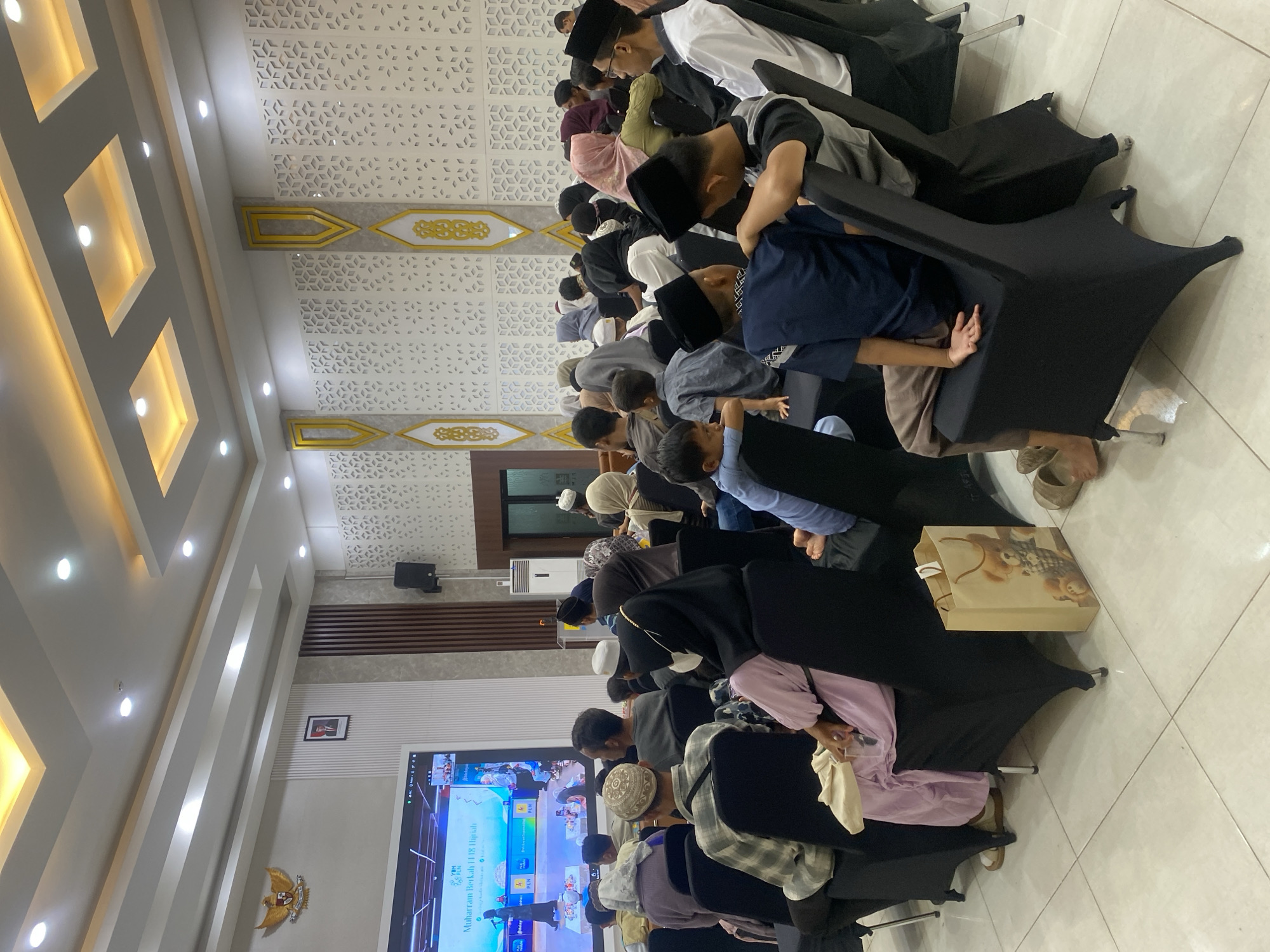 The image depicts an indoor gathering, likely a religious event given the attire of many attendees. A large screen at the front displays text in Indonesian, including "Muharram Berkah 1445 Hijriah," suggesting a commemoration of the Islamic New Year. The room appears to be a hall or conference room, with rows of black-covered chairs occupied by men, women, and children. The attendees are seated and facing the screen. The walls are decorated with geometric patterns and an emblem of Garuda Pancasila, the national emblem of Indonesia, which is visible on the left side of the room. The ceiling features modern lighting fixtures. The floor is tiled. A brown paper gift bag with a teddy bear graphic is visible on the floor next to one of the chairs. The overall atmosphere seems to be one of respectful attendance. While the specific location in Banjarmasin, Indonesia, cannot be determined from the image alone, the presence of the Garuda Pancasila emblem and the Indonesian language on the screen indicates it is within Indonesia.