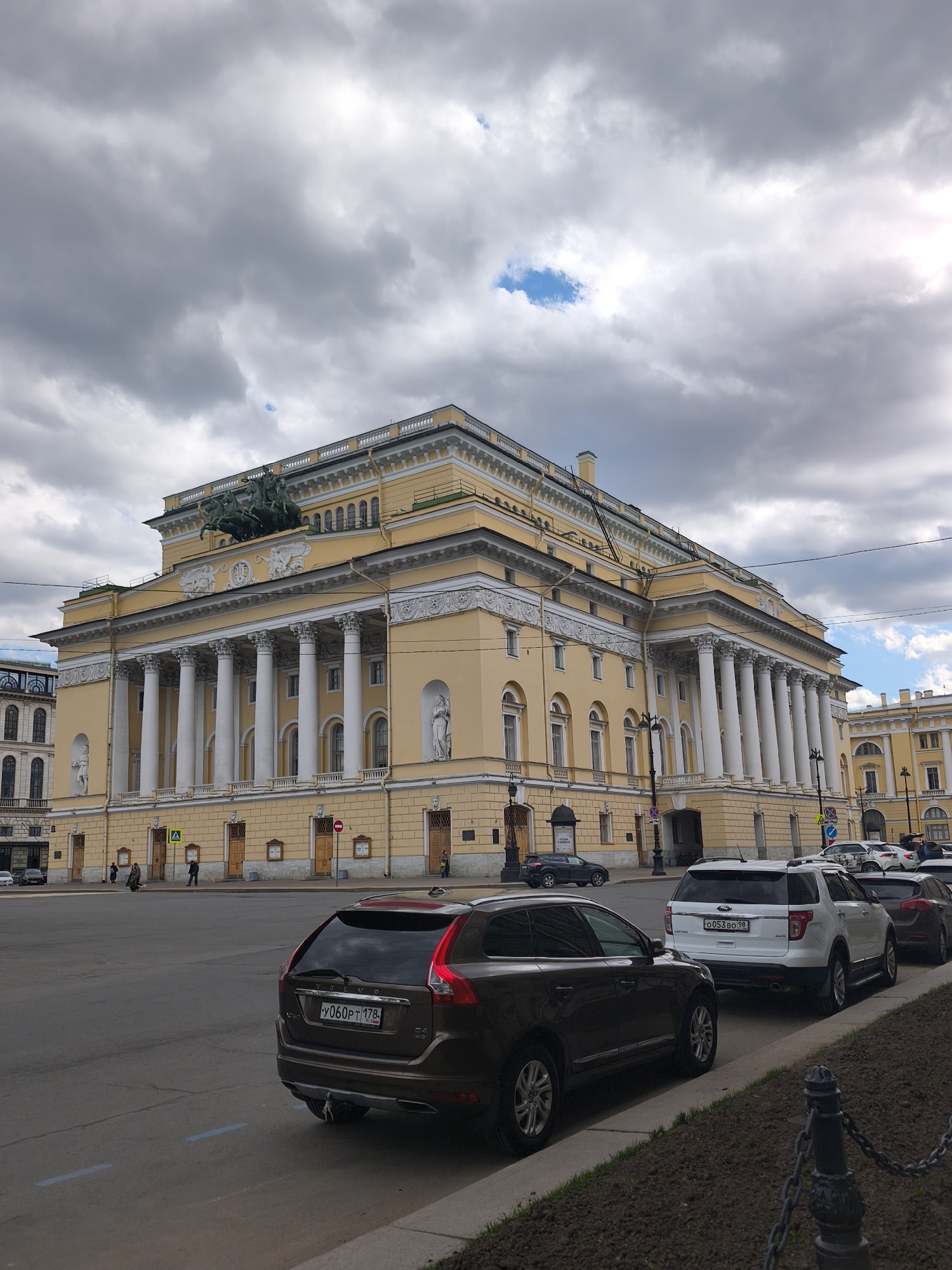 The image captures the Alexandrinsky Theatre in Saint Petersburg, Russia. The theater is a large, pale yellow building with white columns. A sculpture of a chariot pulled by horses is on top of the theater. The sky is overcast with heavy clouds. In the foreground, several cars are parked along the street. A dark brown Volvo SUV with a license plate Y060PT 178 is parked at an angle facing to the right, with a white Ford SUV with license plate O053BO 98 parked behind it. A person is walking across the street near the theater entrance.
